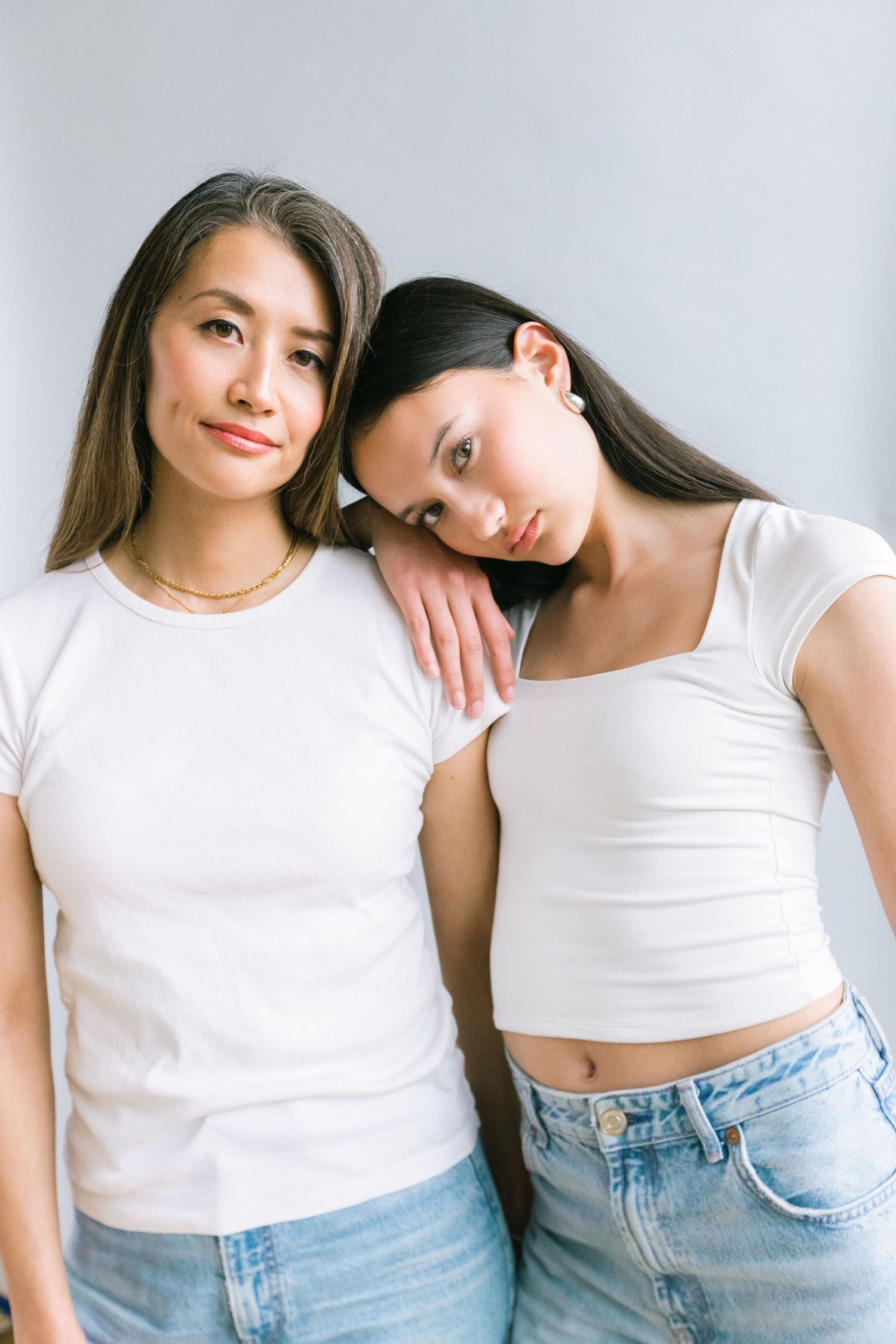 Two young women in white t-shirts and blue jeans posing together against a light gray background.
