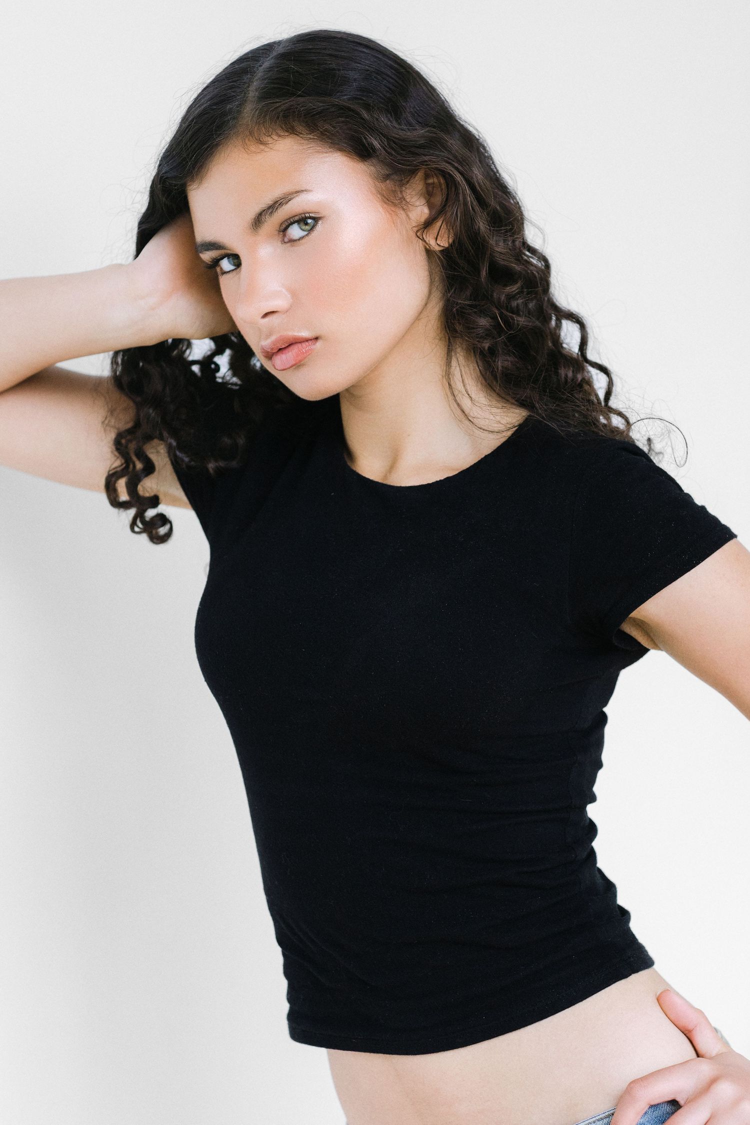 Young woman with curly dark hair wearing a black fitted t-shirt posing confidently against a white background.