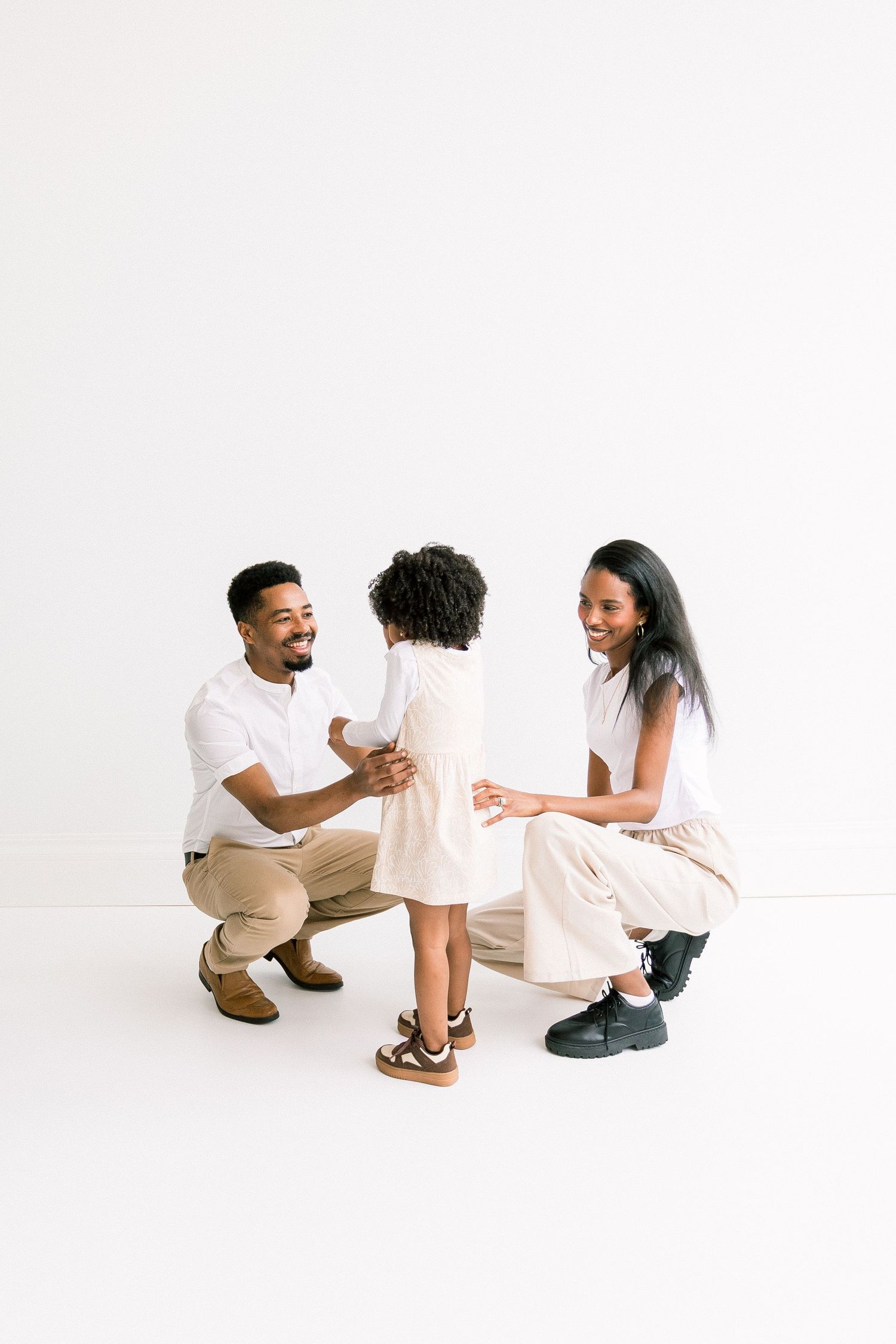 Happy Black family with parents crouching down to interact with their young curly-haired daughter on white background.