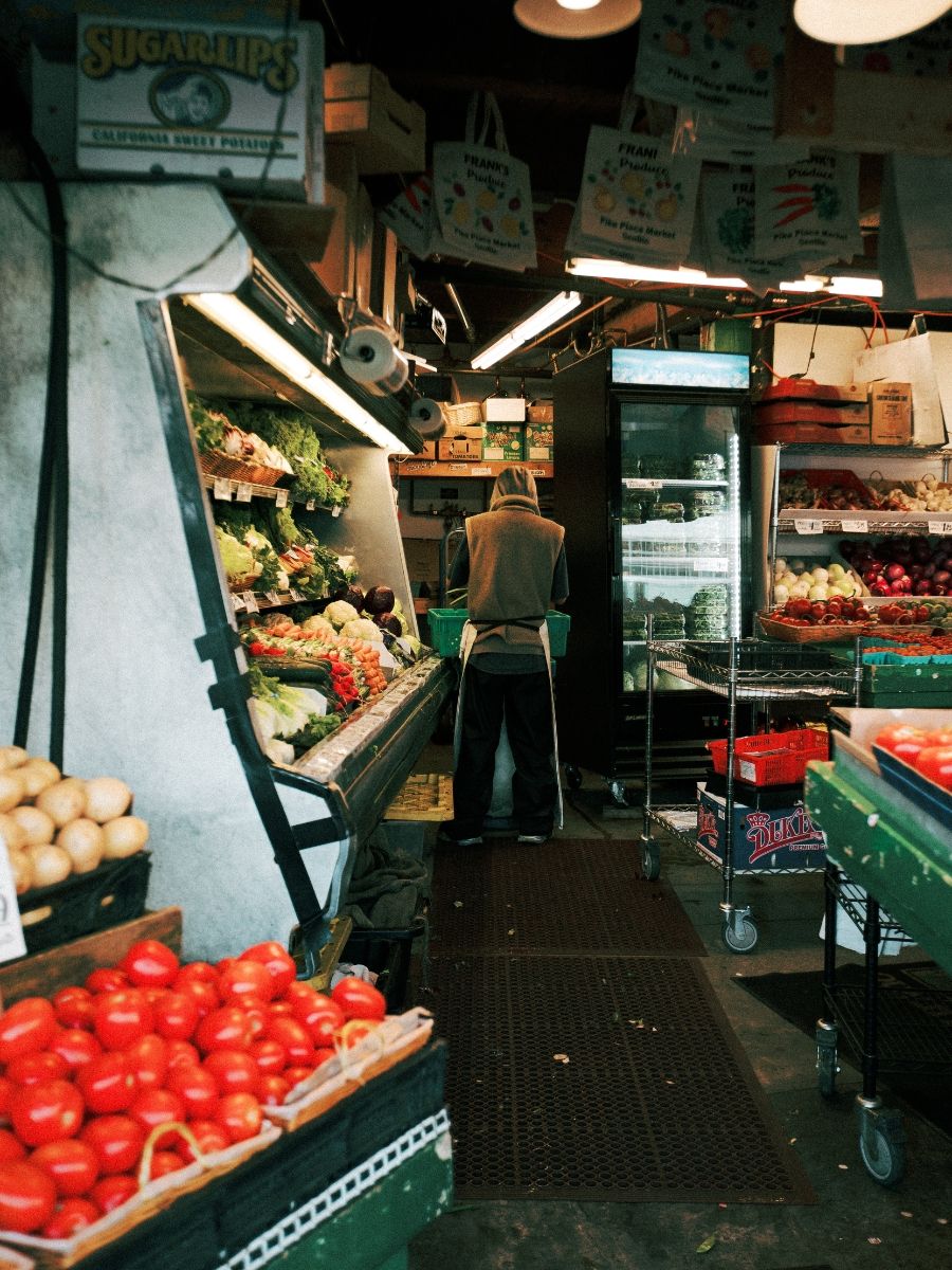 A person shops at a vibrant indoor produce market stall with fresh tomatoes and vegetables on display.