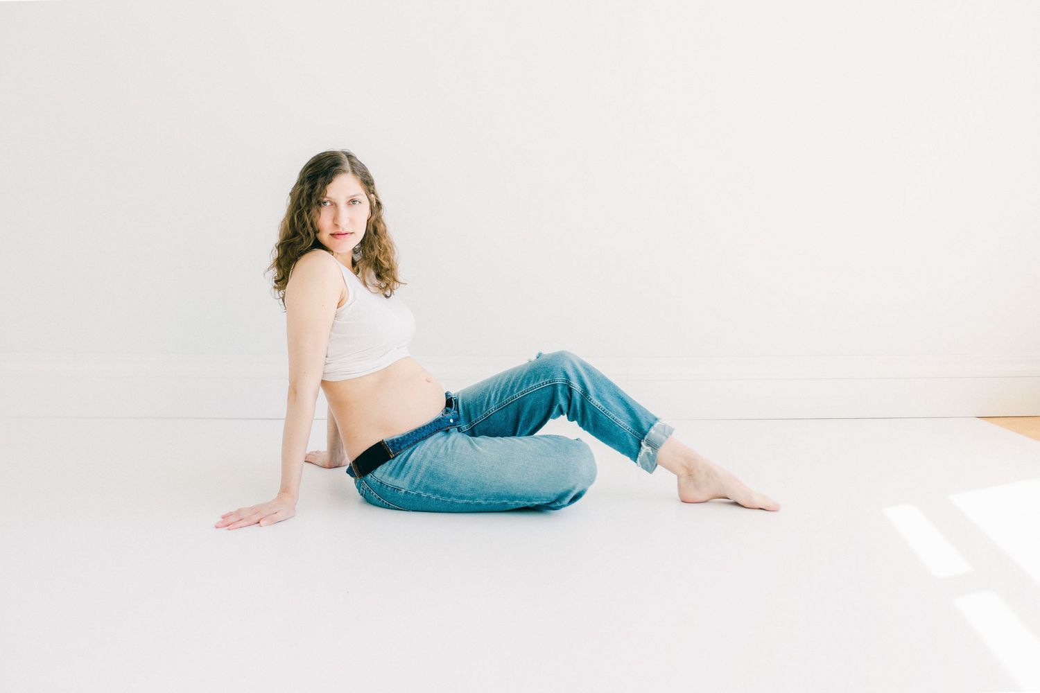 Pregnant woman in white crop top and blue jeans sitting on white studio floor, maternity photo session.