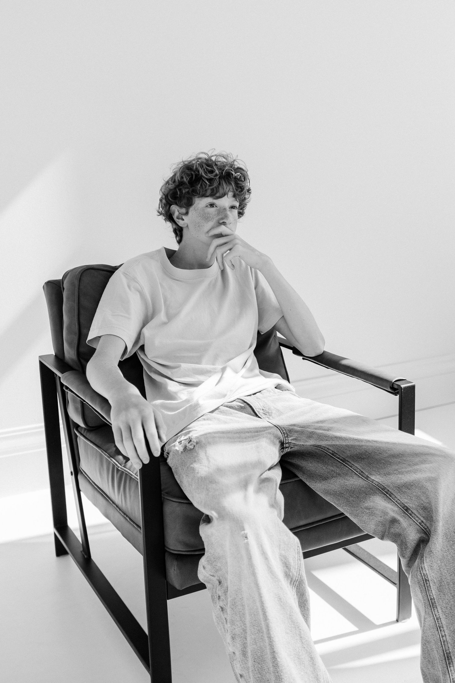 Black and white portrait of a curly-haired young man in a t-shirt and jeans sitting in an armchair.