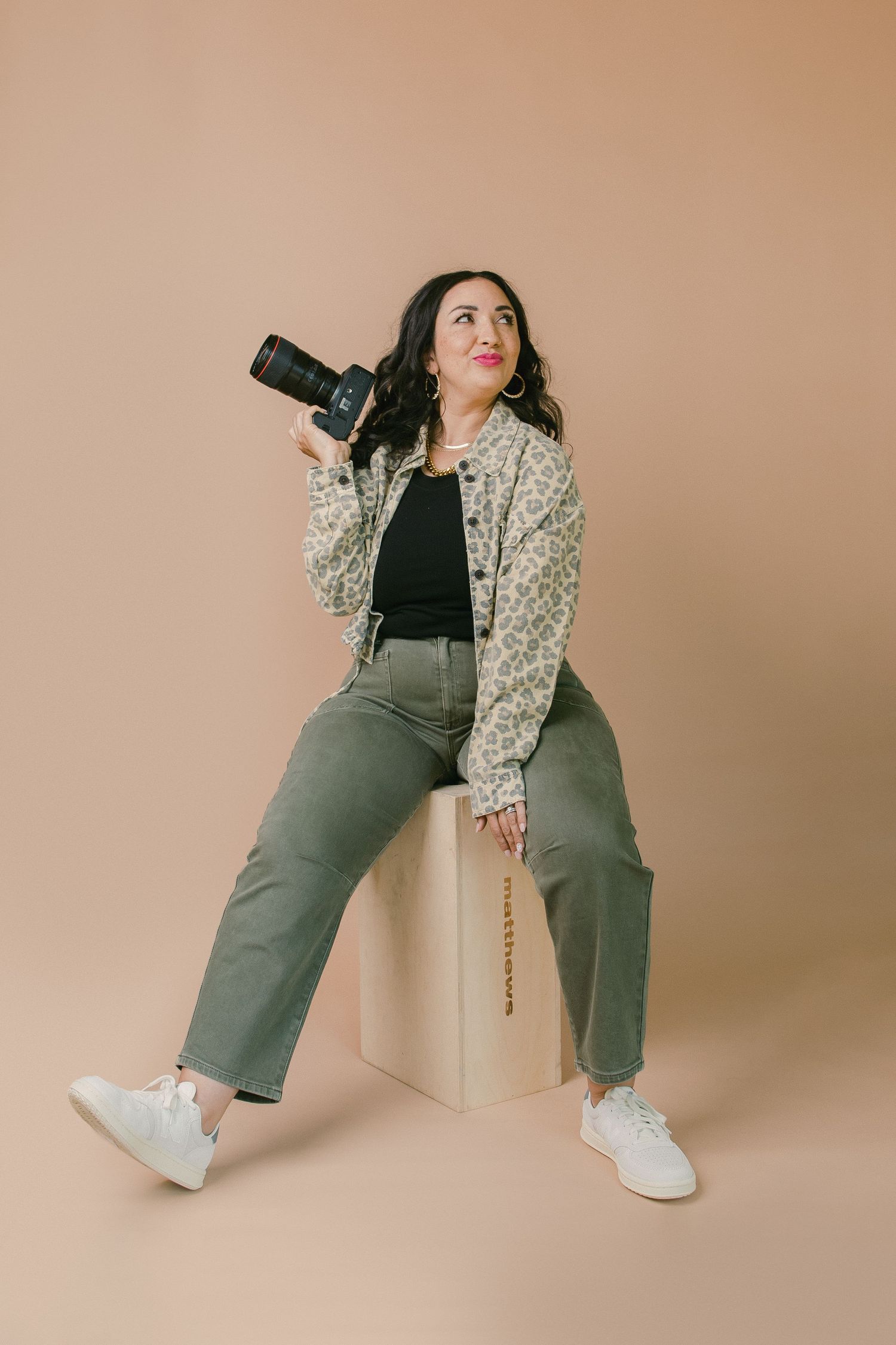 Woman sitting on a white box holding a camera, wearing a floral jacket, green pants, and white sneakers on a beige background.