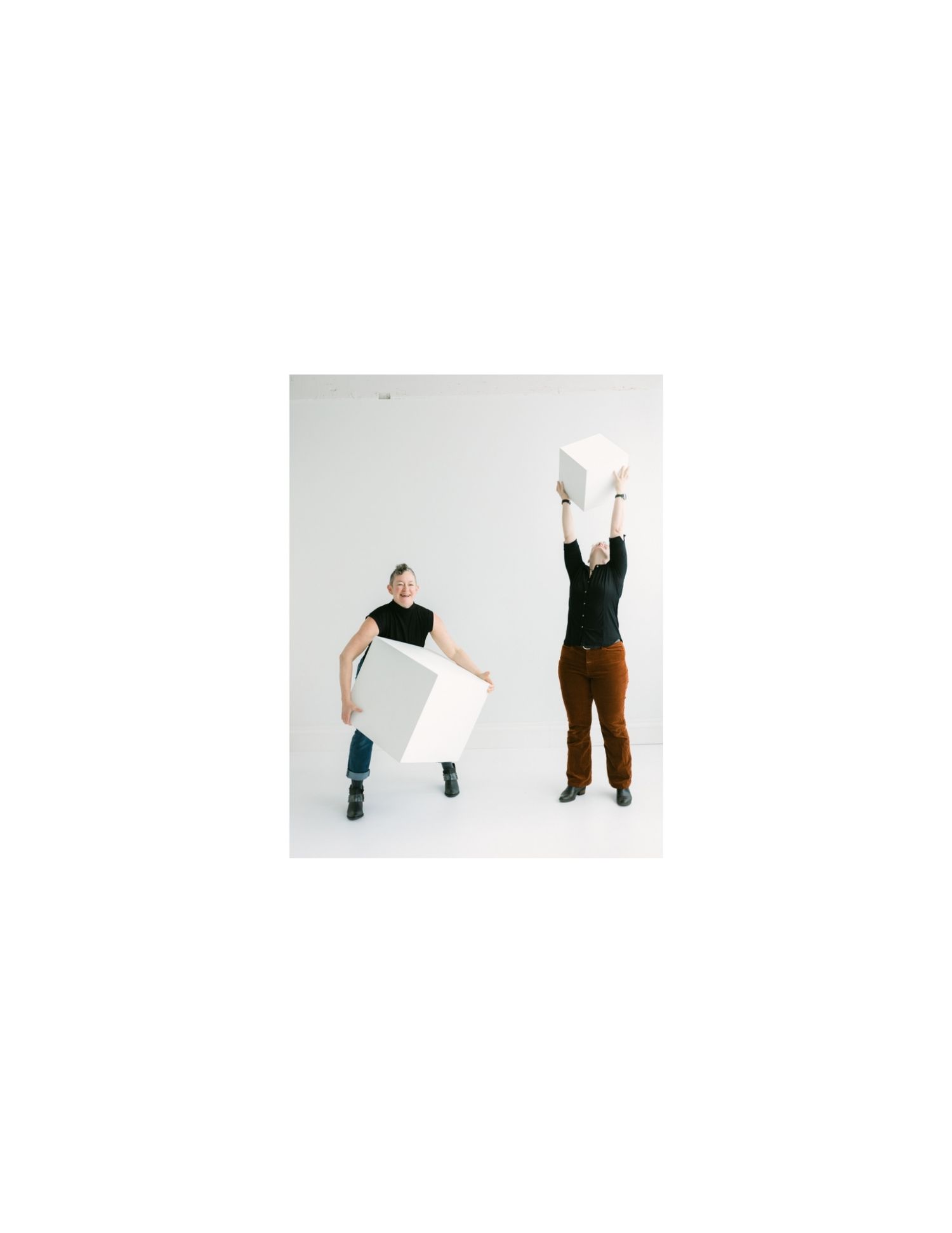 Two people holding large white cube shapes in a studio, one lifting it overhead and one holding it at their side.