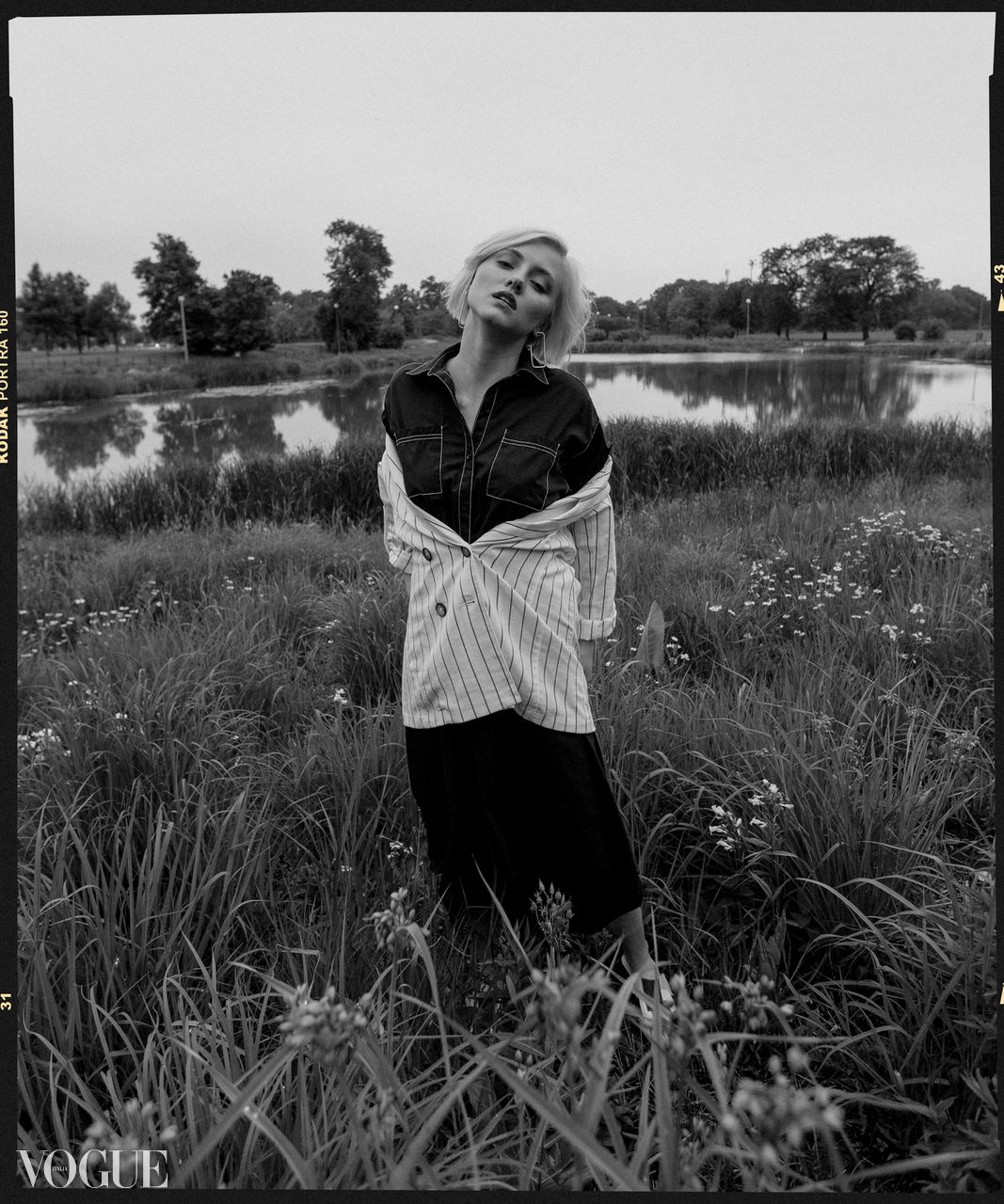 Black and white Vogue editorial of a blonde woman posing in a field near a pond at dusk.