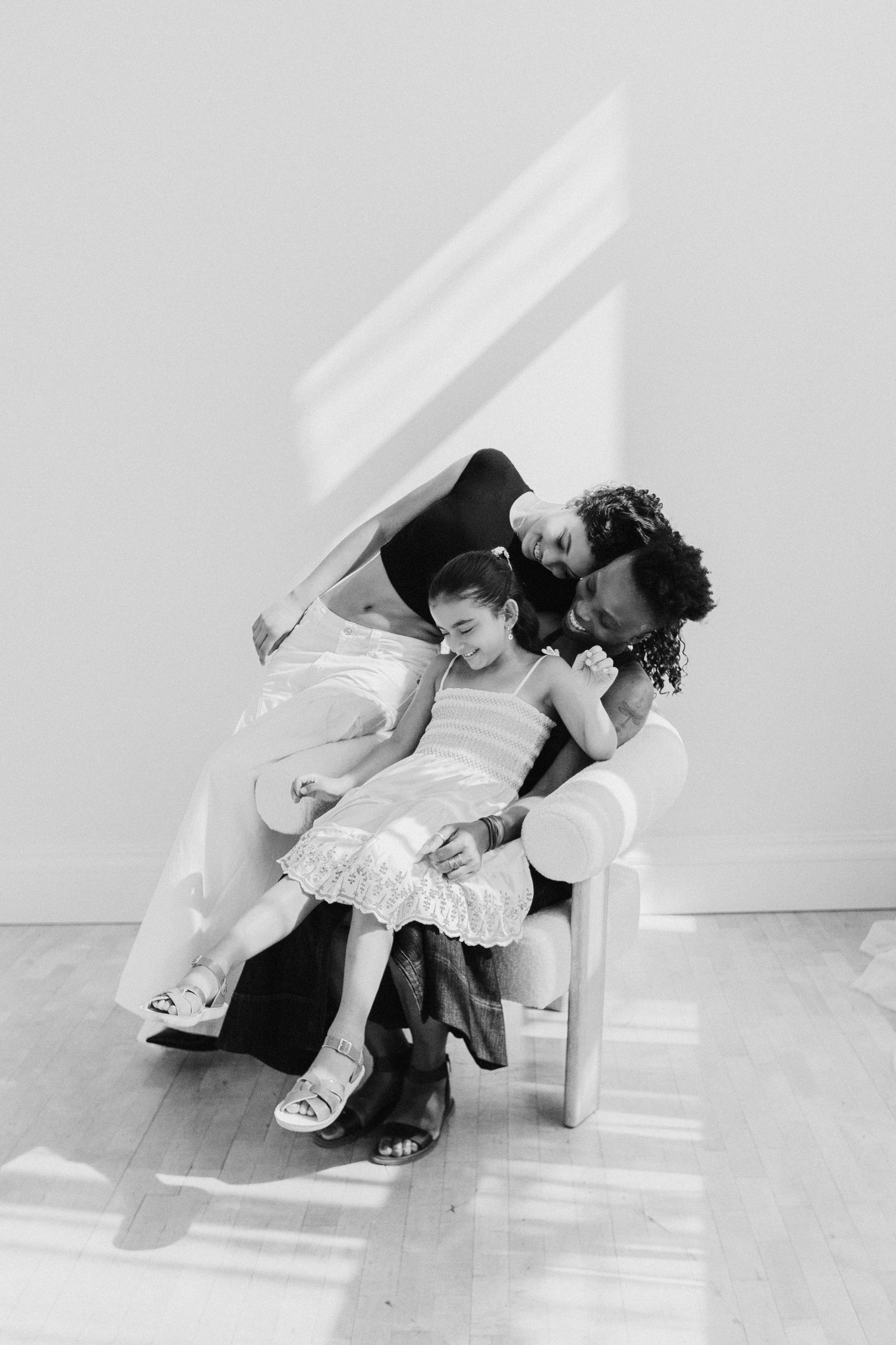 Black and white photo of two parents lovingly embracing their young daughter seated in a white chair indoors.