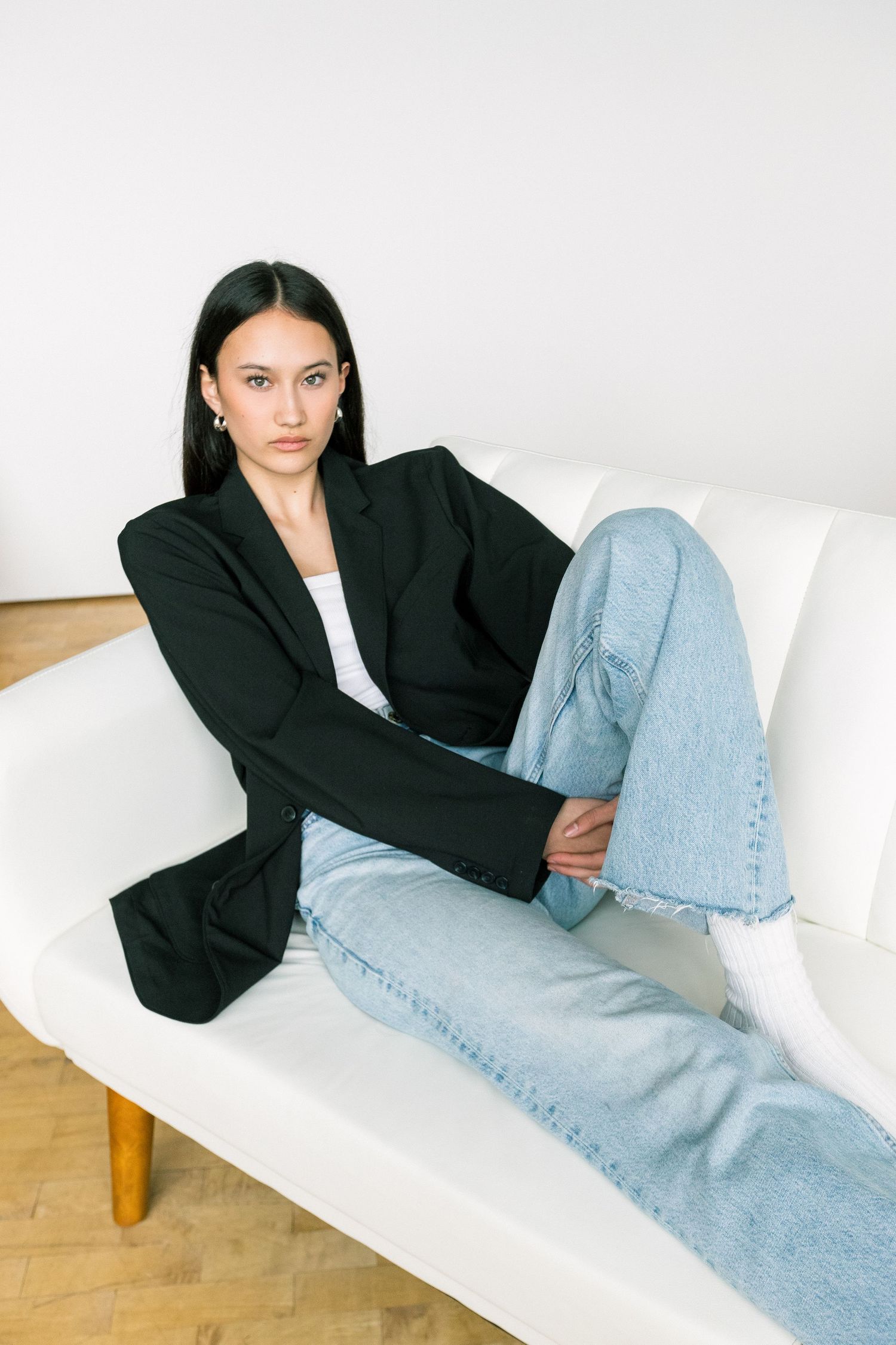 Woman in black blazer and light blue jeans sitting relaxed on white sofa against white wall.