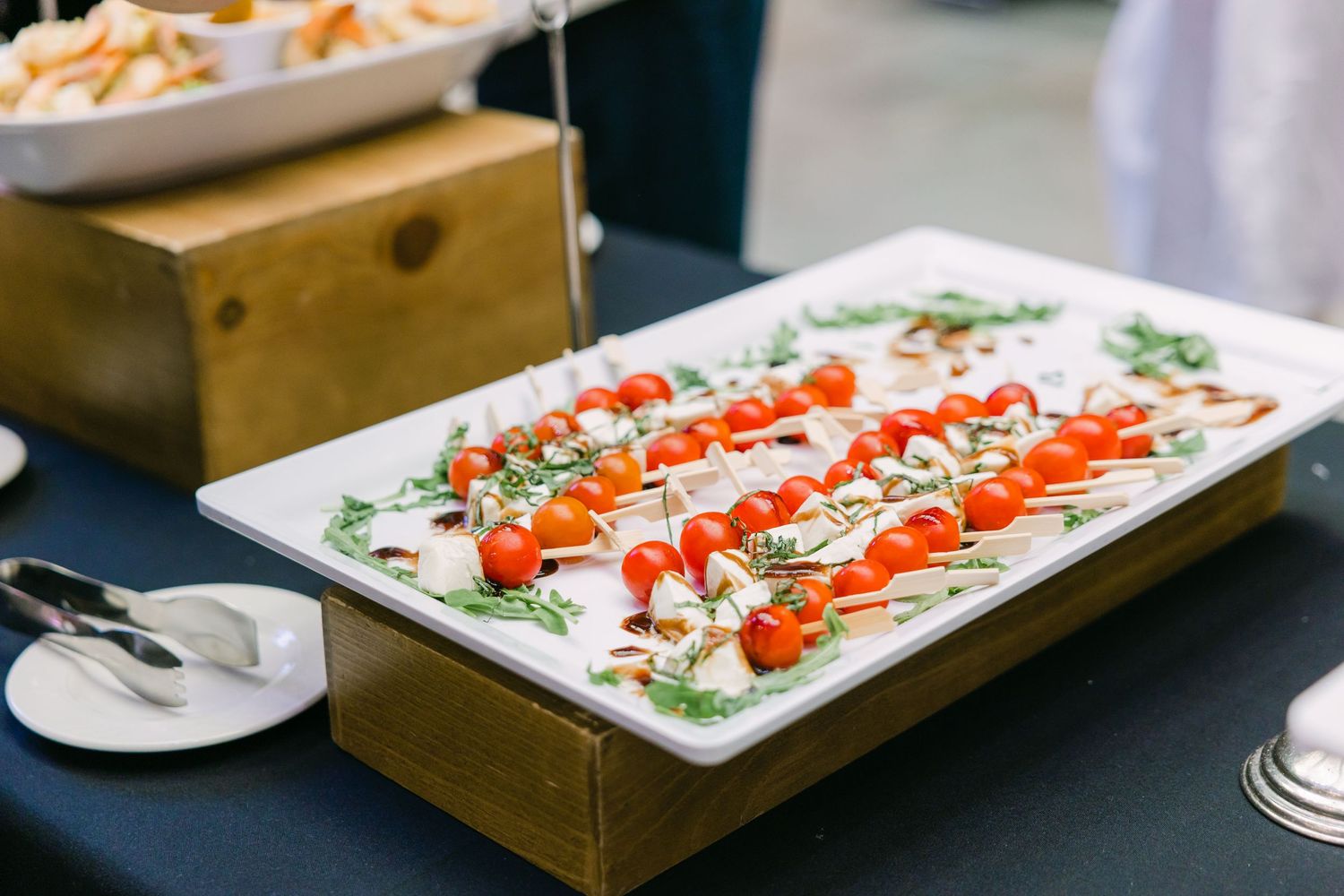 Elegant caprese skewers with cherry tomatoes and fresh mozzarella on white rectangular platter at catered event.