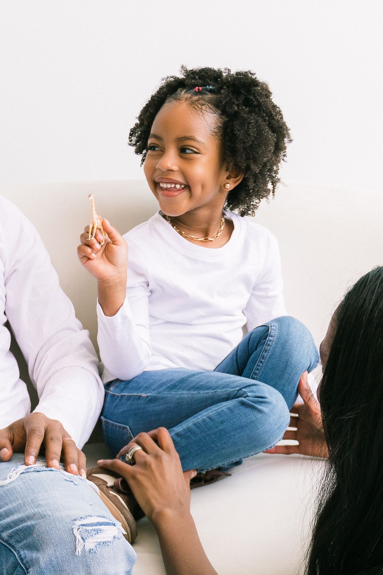 A joyful young Black girl sits cross-legged on a sofa, smiling and holding a pencil, flanked by adults.