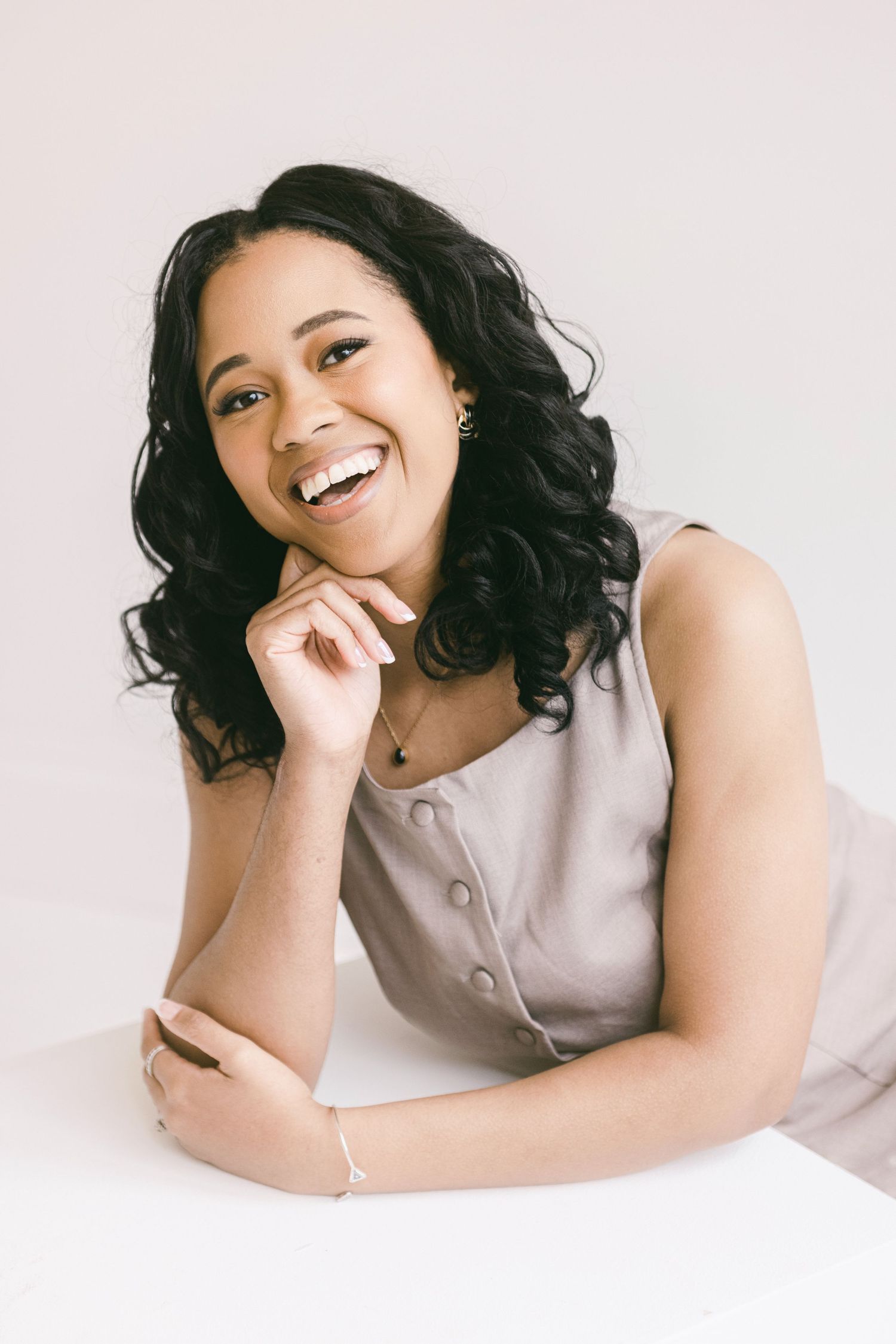 Smiling young woman with curly hair in a gray sleeveless top, posing confidently against a white background.