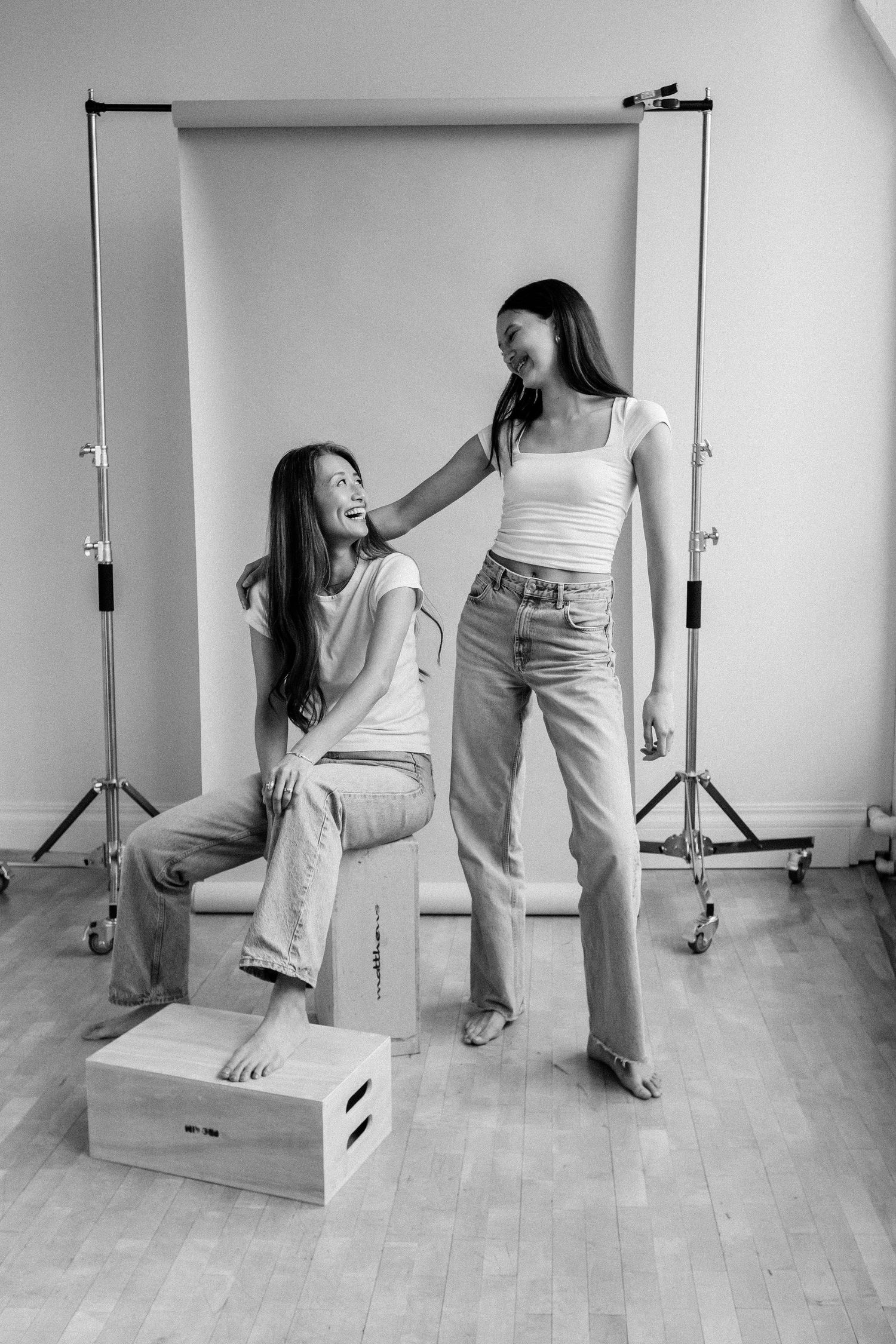 Two teenage girls laugh together in a photography studio with backdrop and lighting equipment.