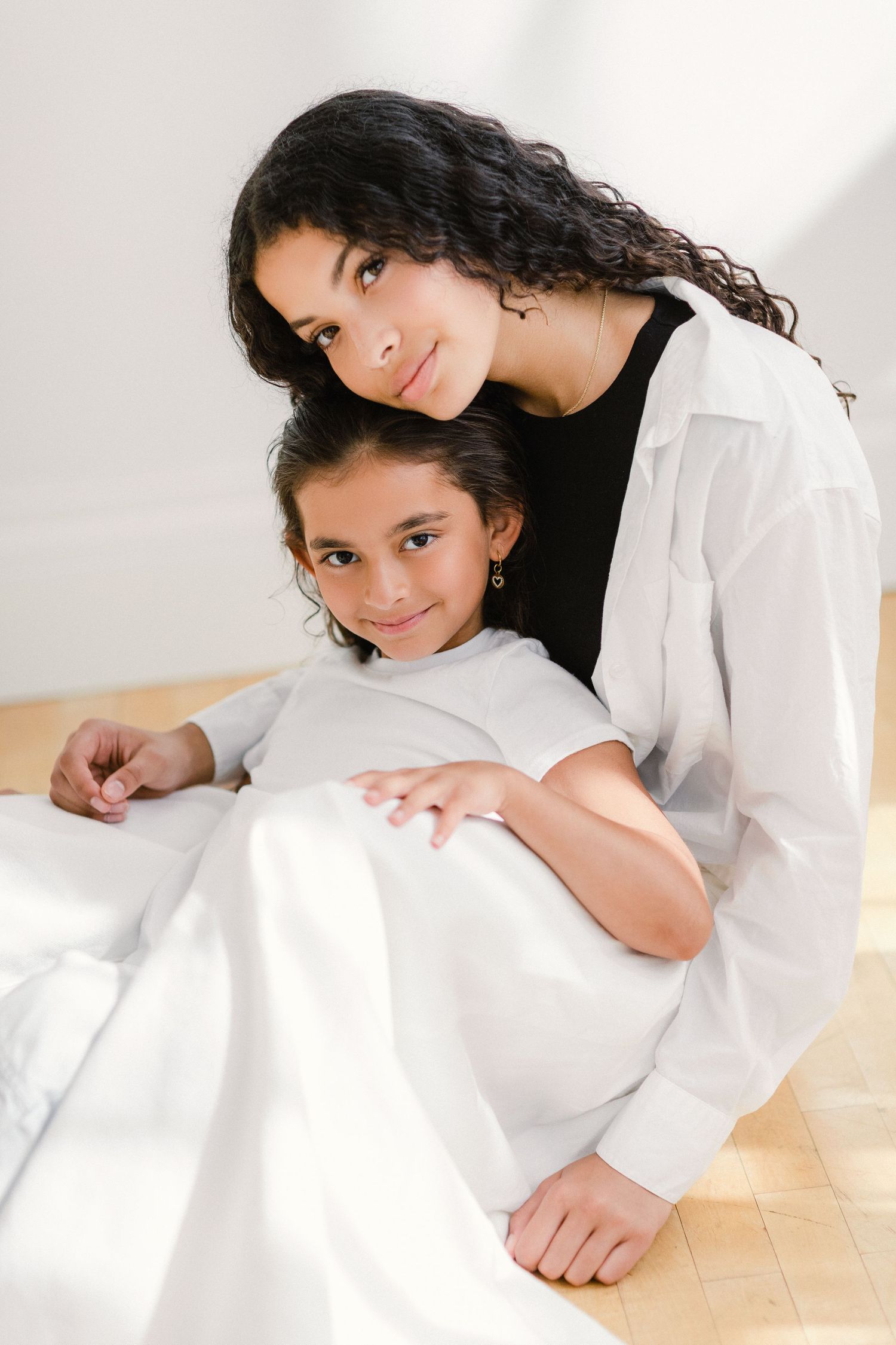 A mother and young daughter sitting together on the floor, smiling warmly at the camera indoors.