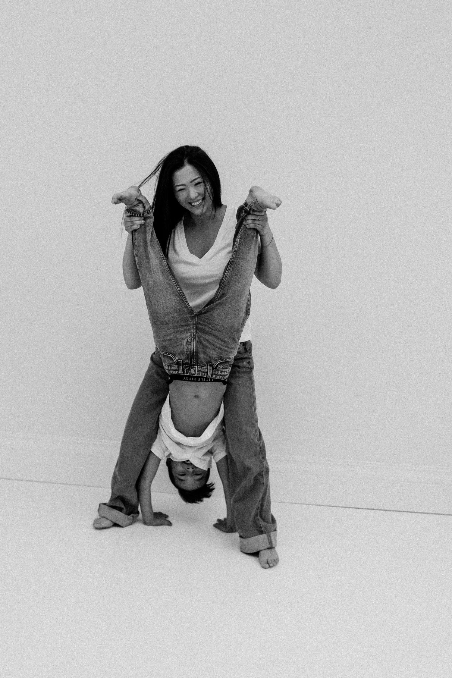 Woman smiling while holding a child upside down playfully, both wearing jeans, black and white photo.