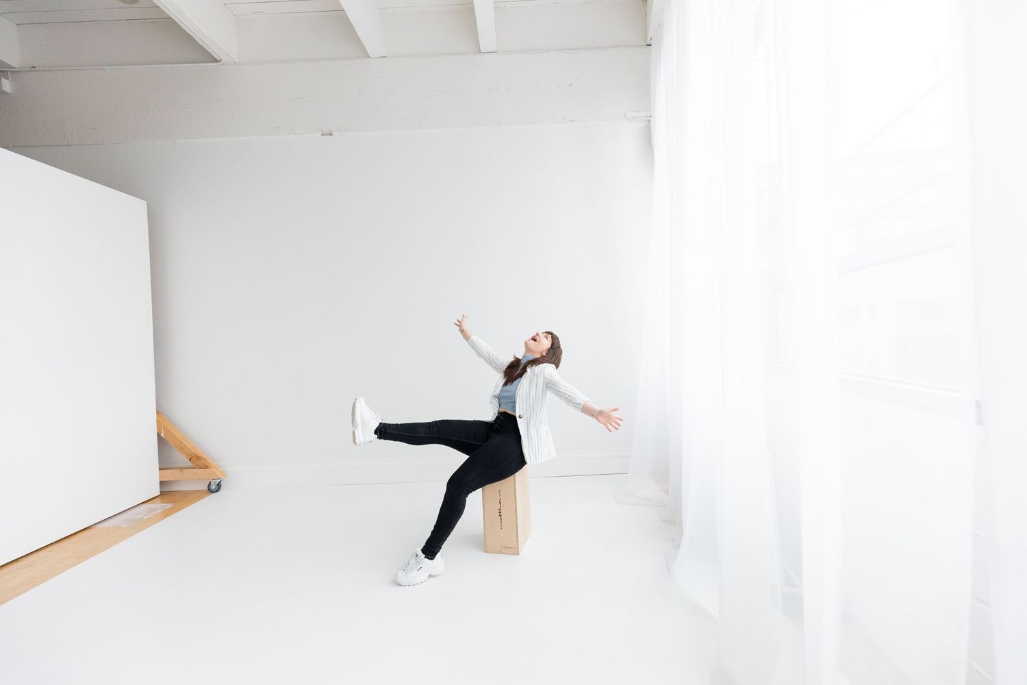 Woman in black pants and gray top poses dynamically on a wooden bench in a bright white studio space.