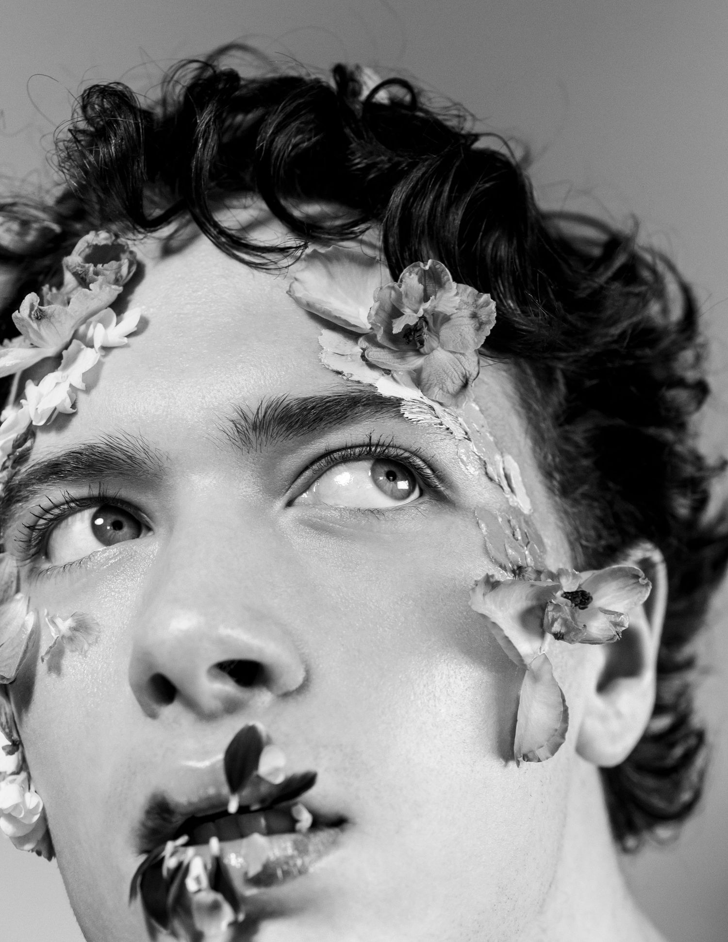 Black and white portrait of a young man with curly hair adorned with delicate flowers on face and hair.