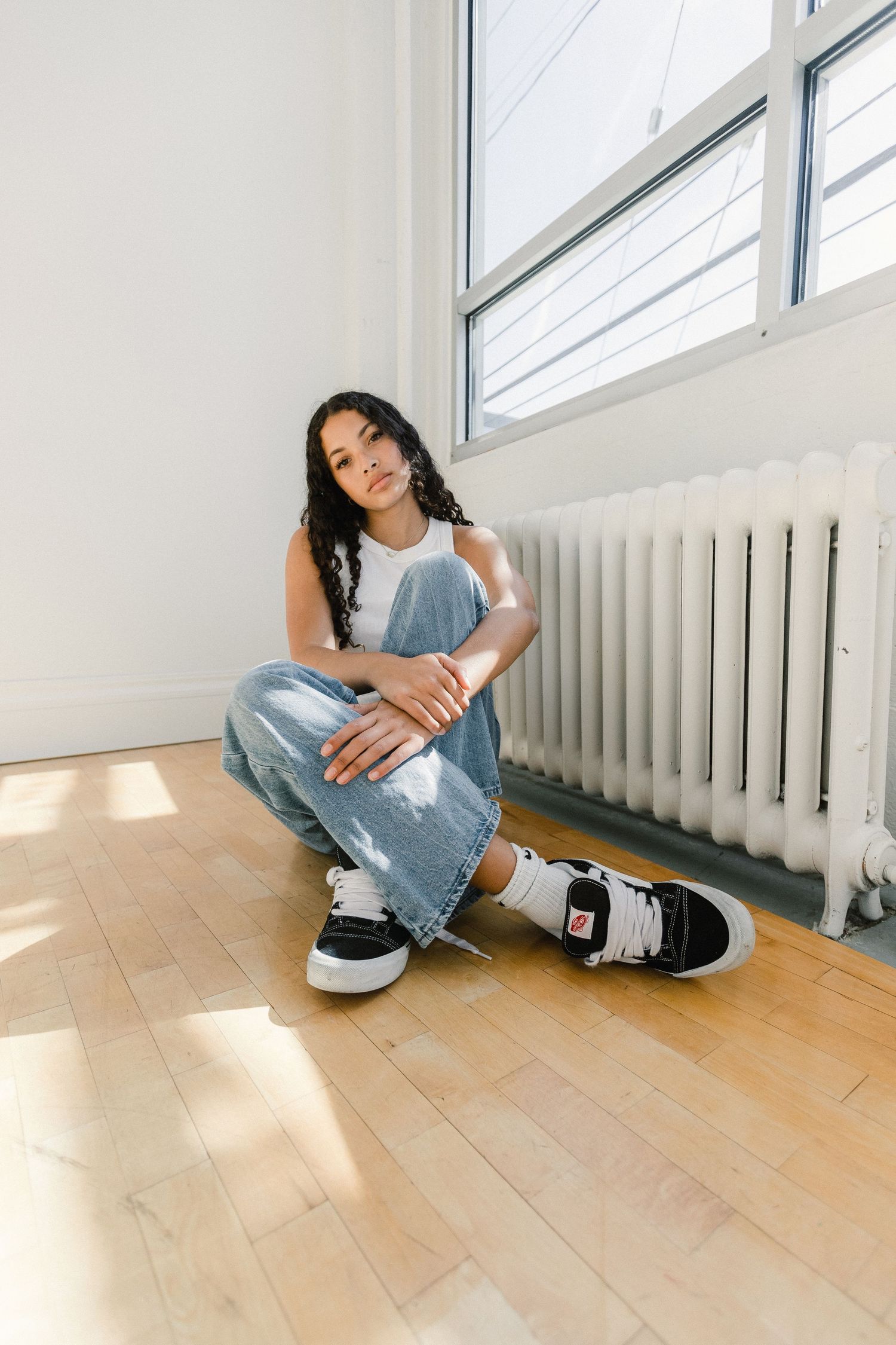 Young woman in jeans and Vans sneakers sitting on hardwood floor near window.