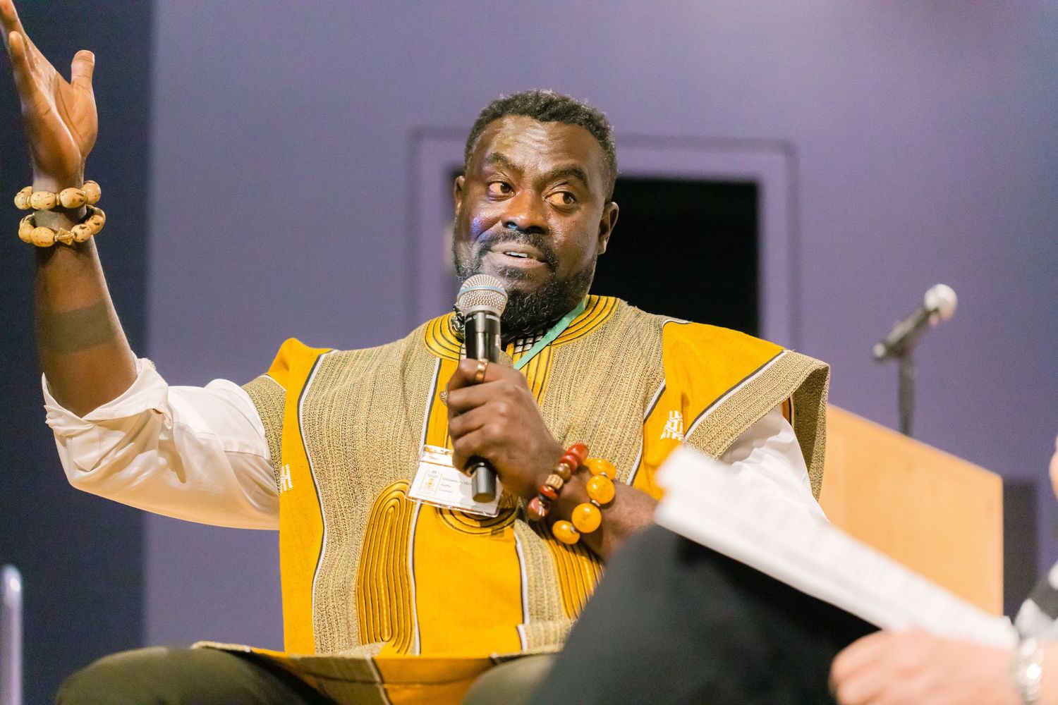 Energetic male speaker in yellow African attire gestures while holding microphone at conference.