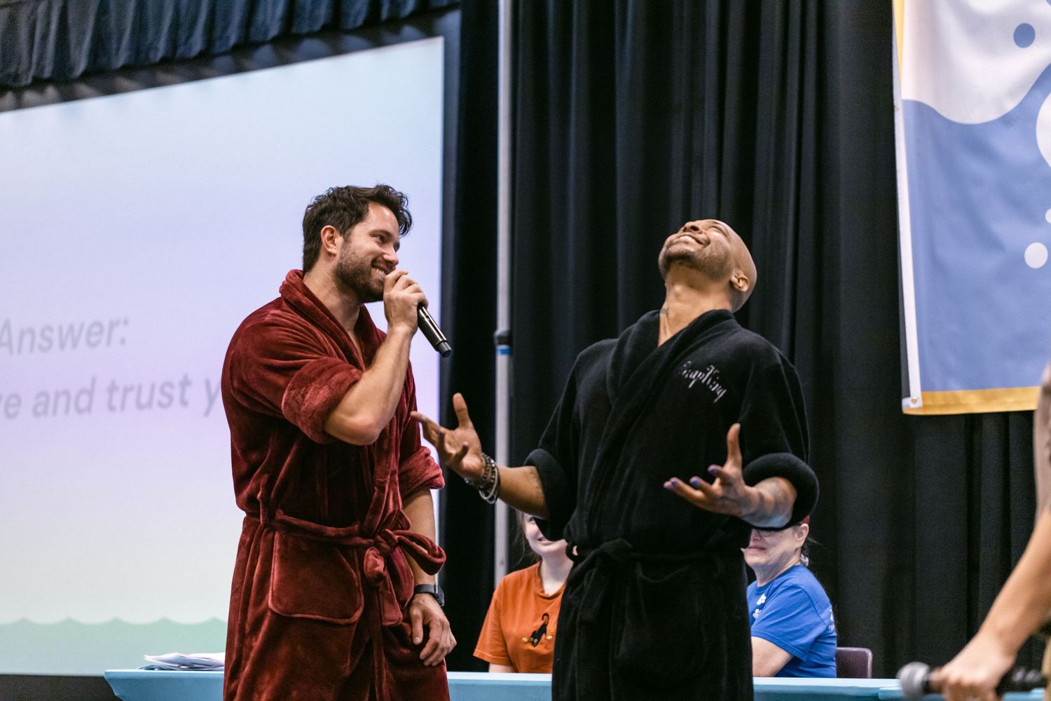 Two men performing on stage with a microphone at an indoor event with a presentation screen behind them.