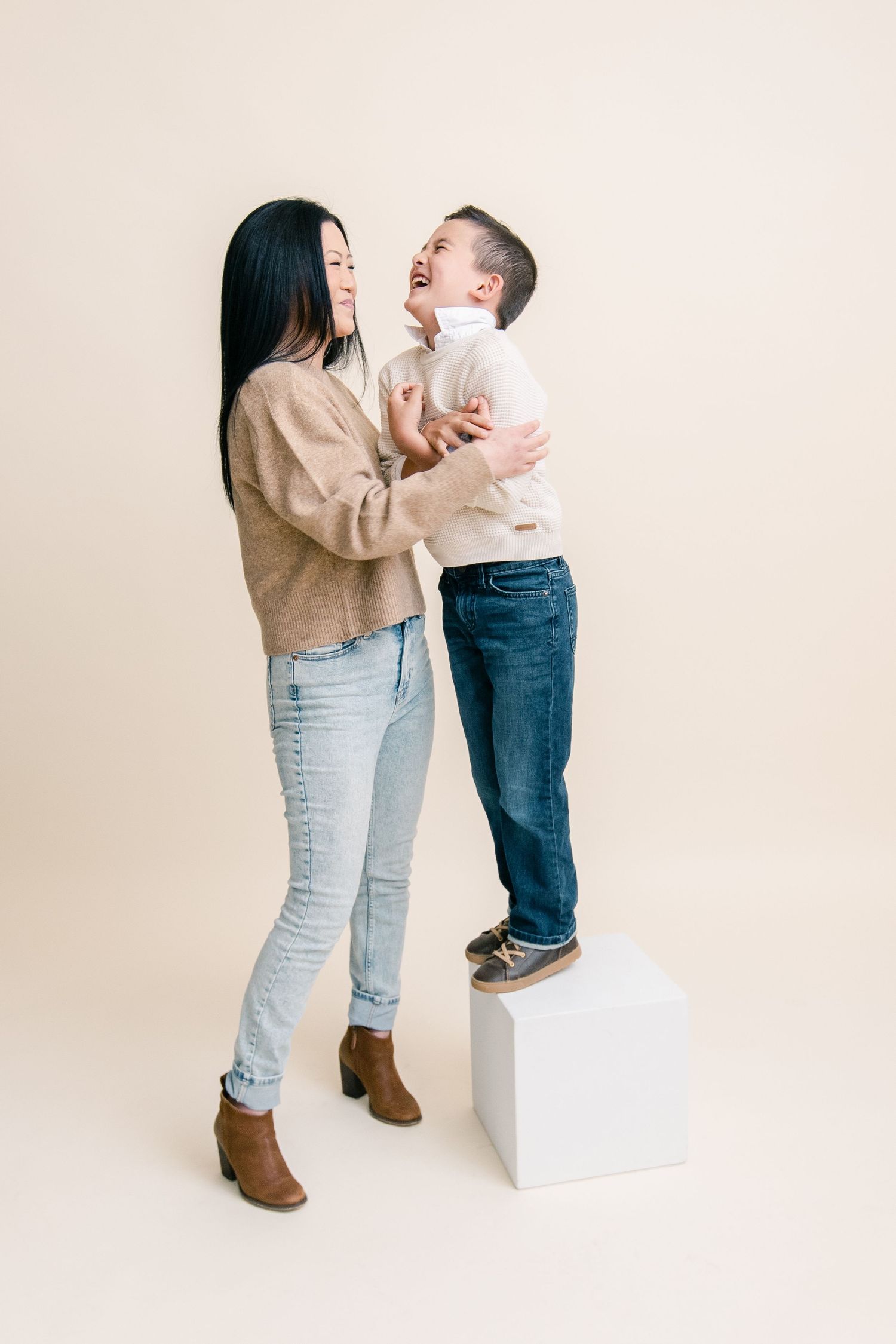 A woman and young boy laughing together in a studio, both wearing jeans and sweaters on a beige background.