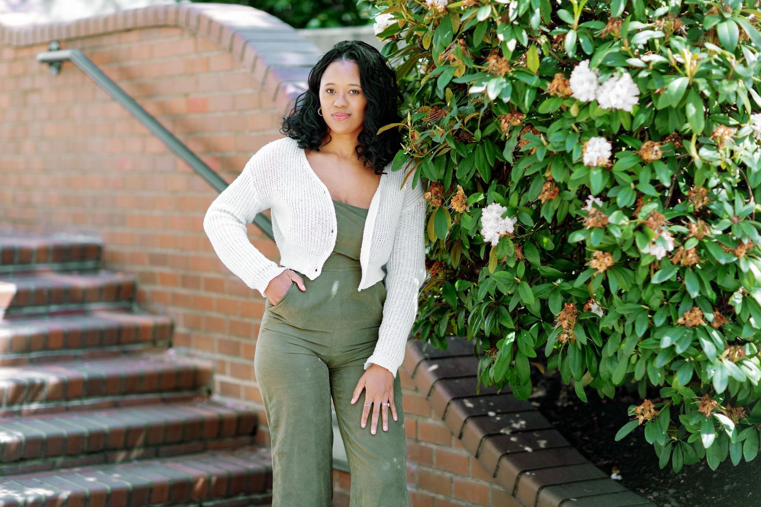 Confident woman in white cardigan and olive green pants posing outdoors near brick steps and flowers.
