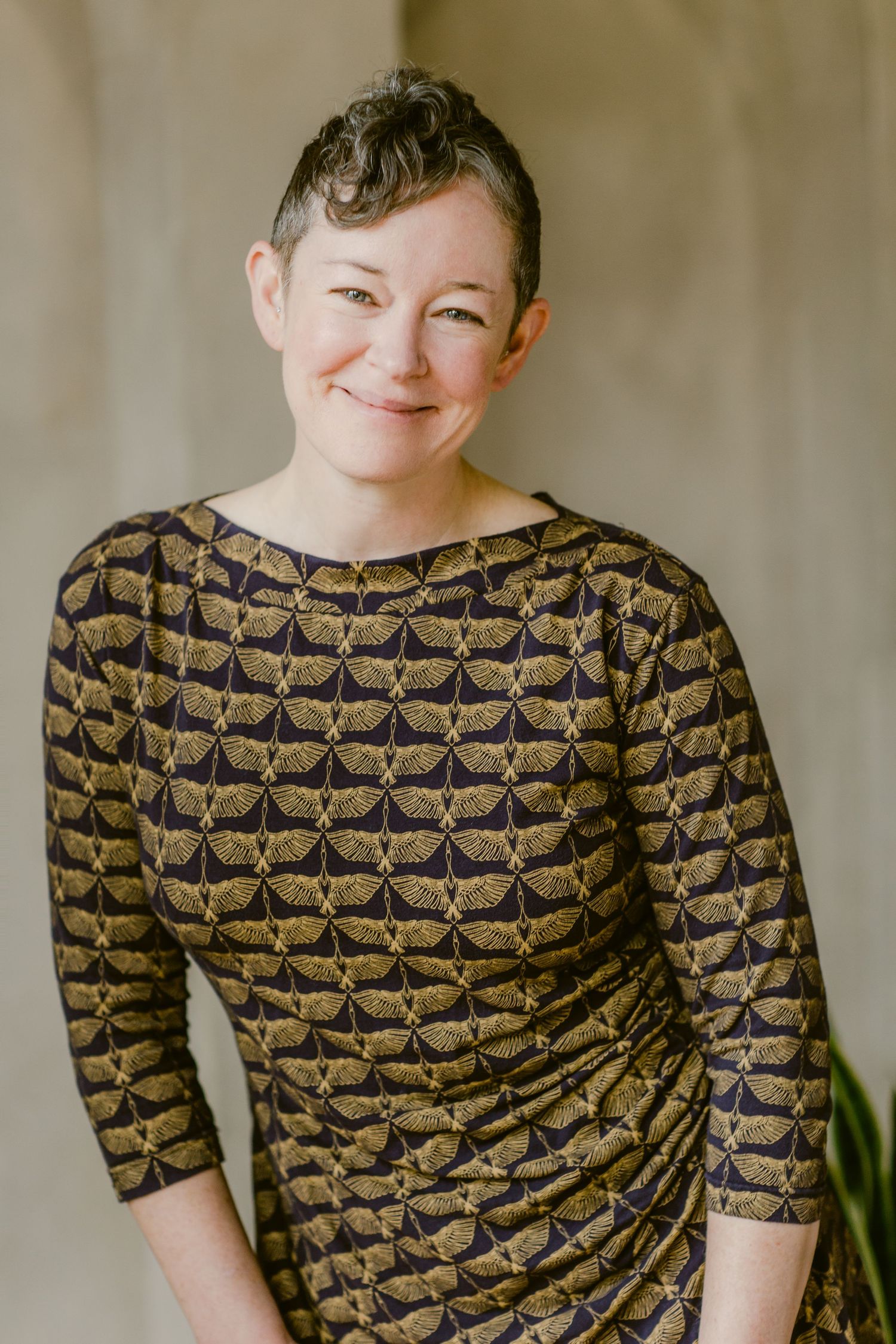 Woman smiling in a gold and black patterned dress, posing confidently indoors for a professional portrait.