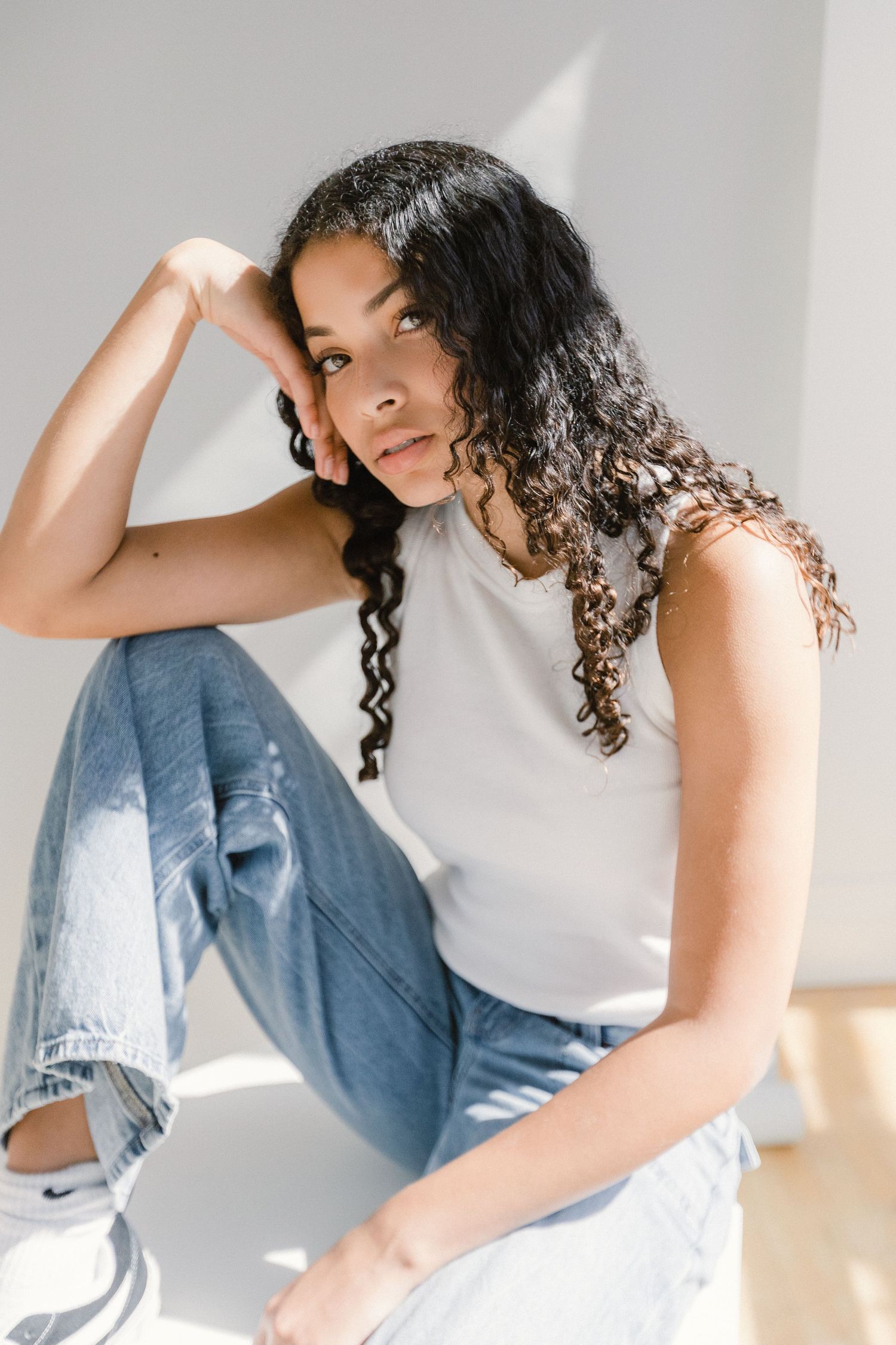 Young woman with curly hair in white tank and ripped jeans sits on floor in natural sunlight.