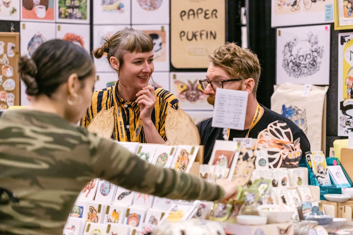 Two vendors smile at a customer at a Paper Puffin art fair booth displaying colorful prints.