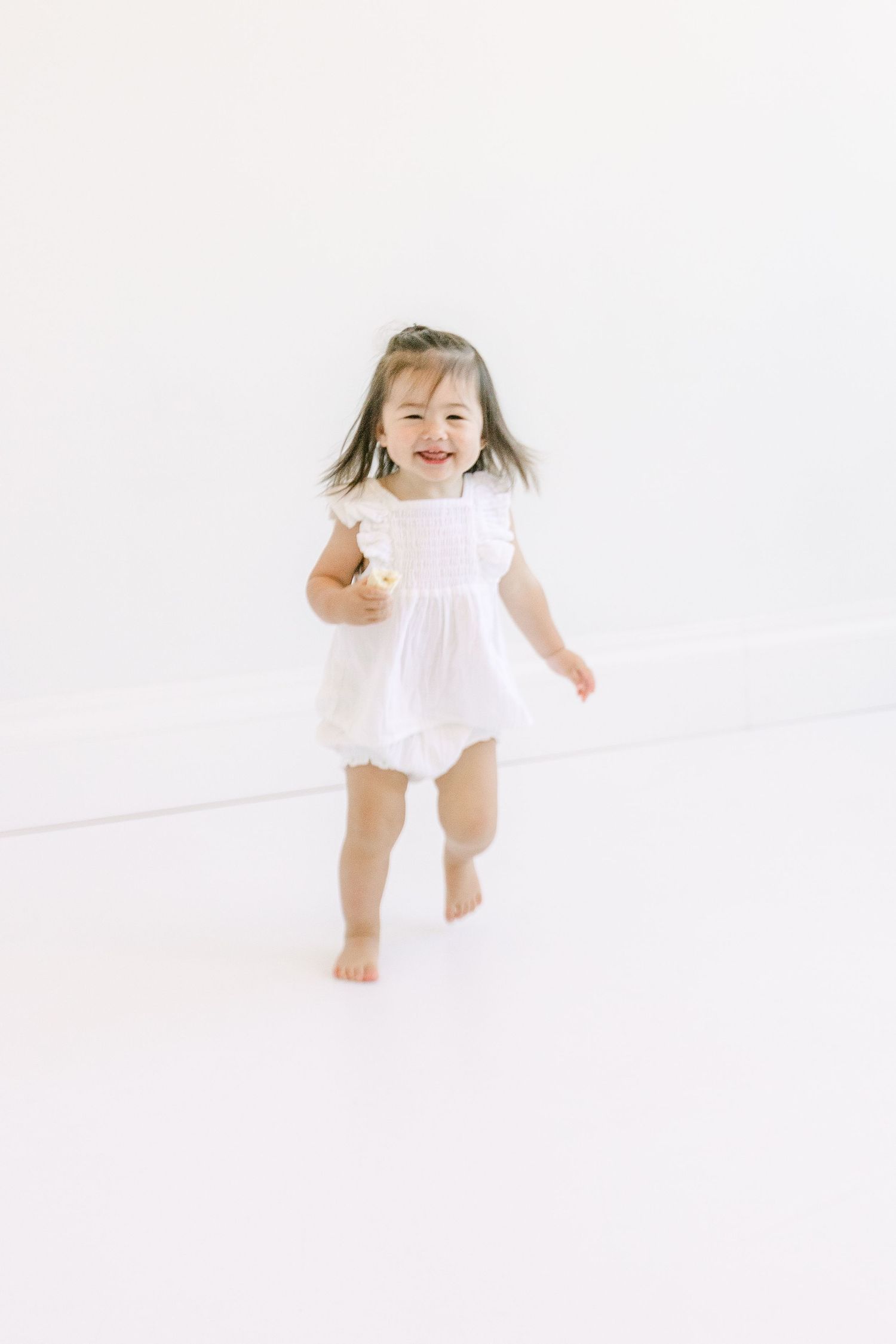 Smiling toddler girl in white dress running barefoot against a bright white background.