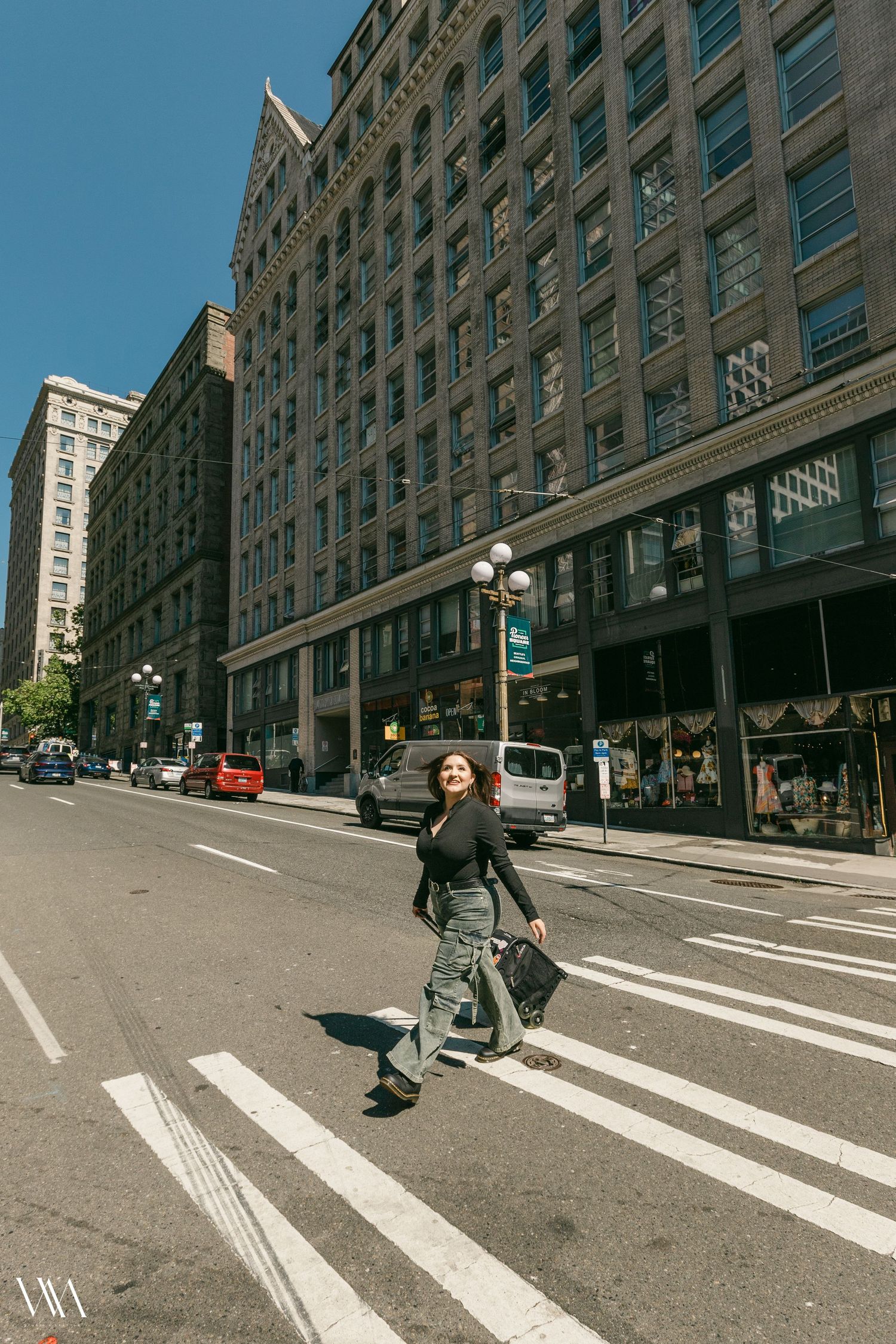 Woman in black top and camo pants crossing a downtown city street crosswalk with large historic buildings behind her.