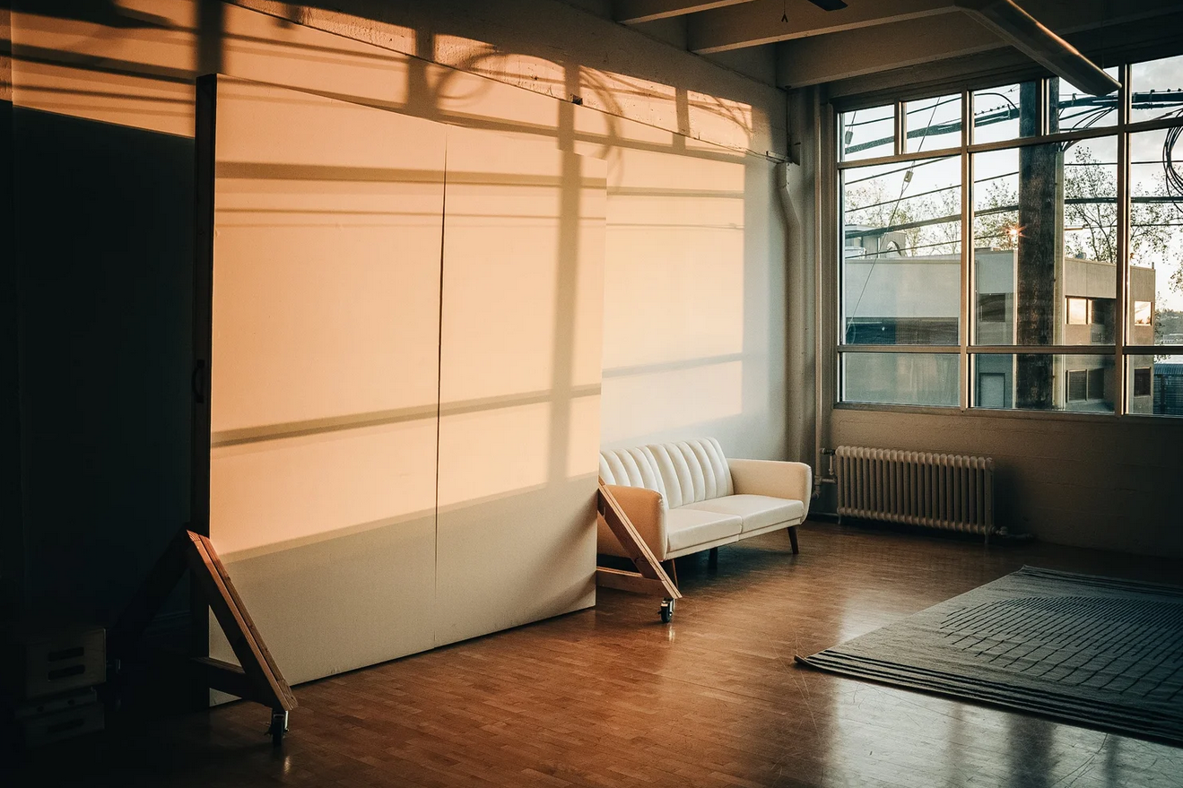 Minimalist studio space with white sofa, wooden floors, large windows, and warm sunlight casting shadows on walls.