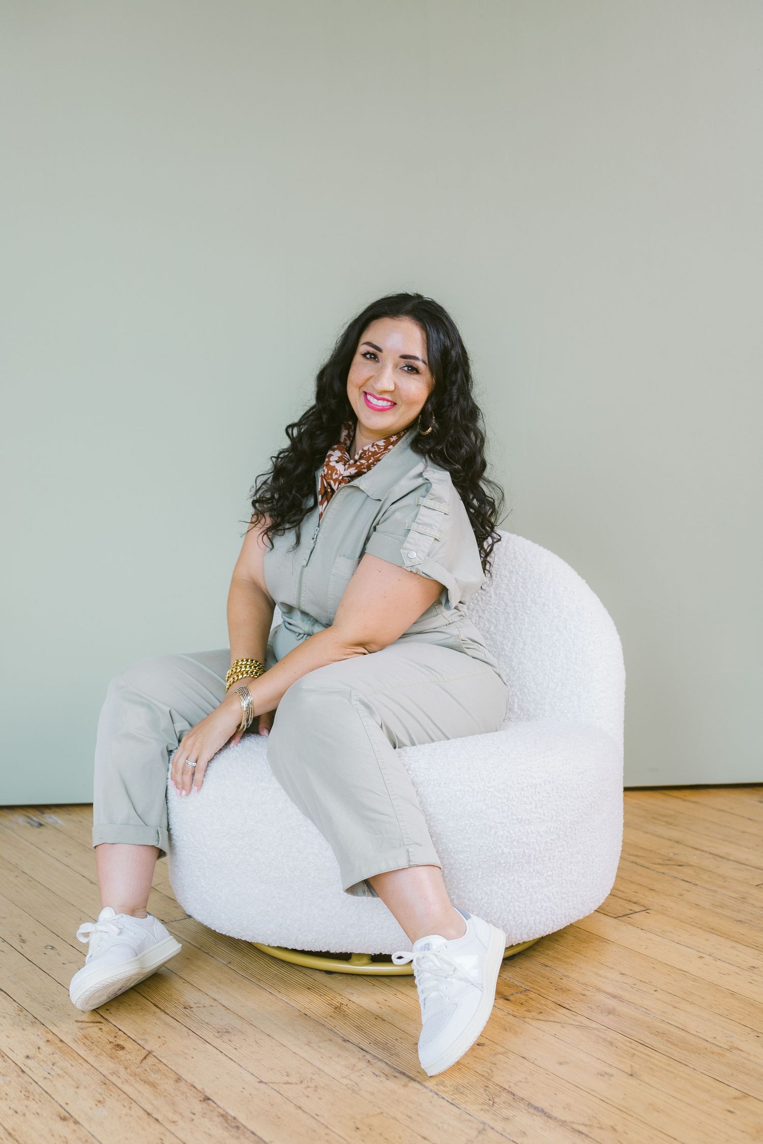 Woman in sage green jumpsuit sitting in white boucle chair, smiling, wearing gold jewelry and white sneakers.
