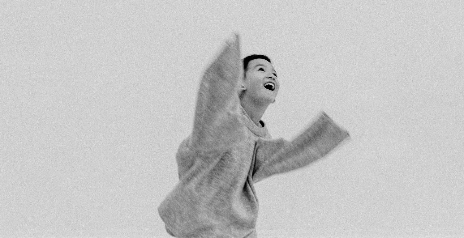 A boy leaps with arms outstretched and head thrown back in pure joy against a light gray background.