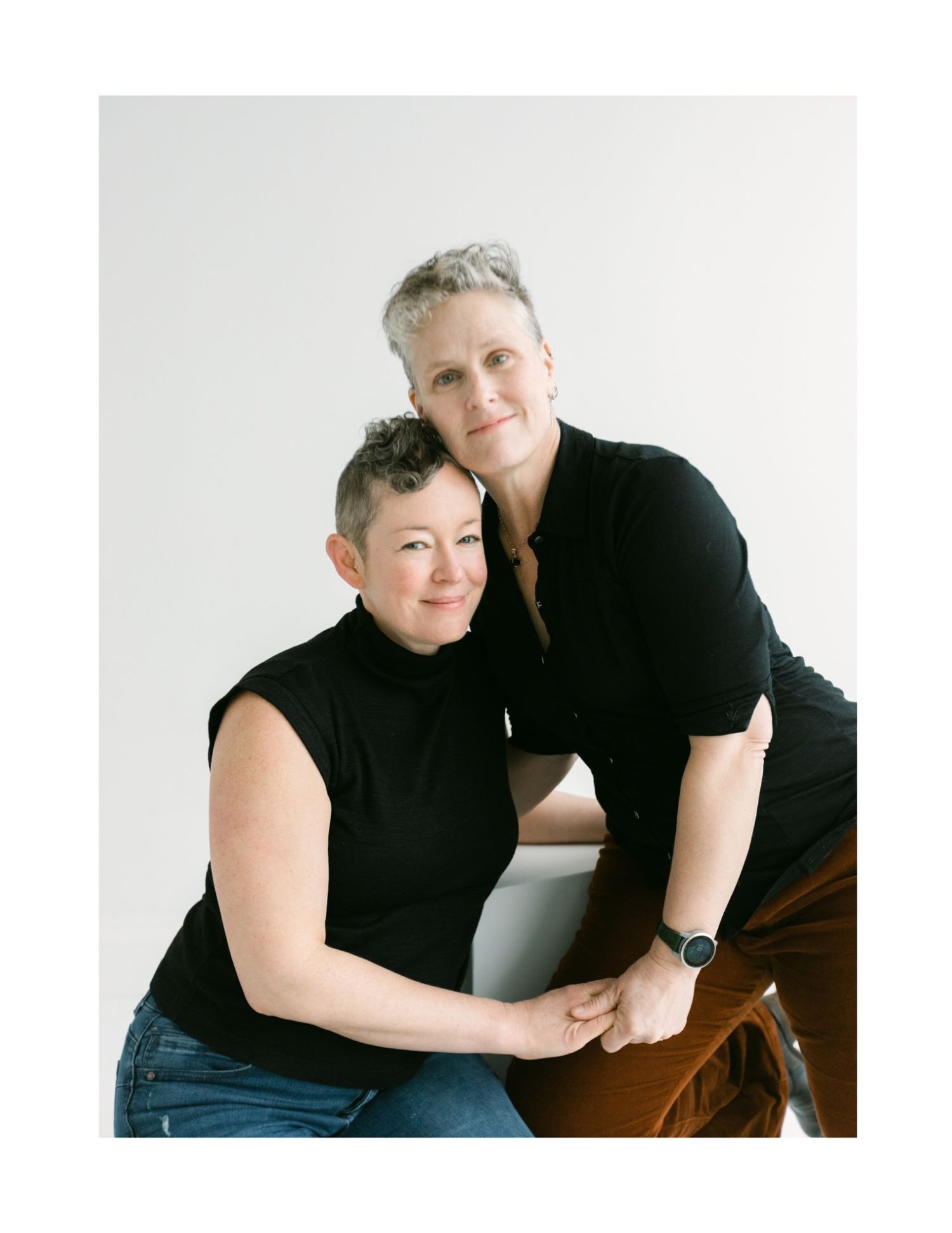 Two women with short gray hair embrace warmly, both wearing black tops, in a bright studio portrait.