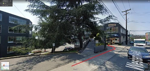 Street view of urban buildings with trees, a red arrow marking a driveway entrance, and power lines against a clear sky.