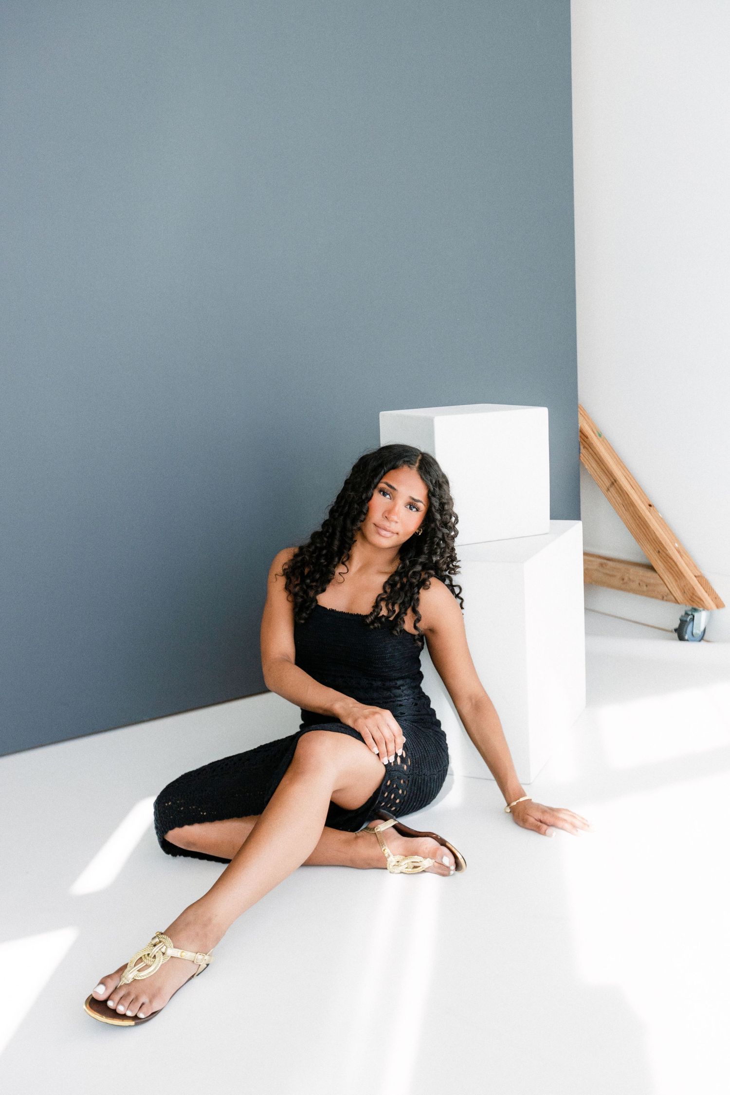 A young woman with curly hair sits on a white floor in a black dress and gold sandals against a gray wall.