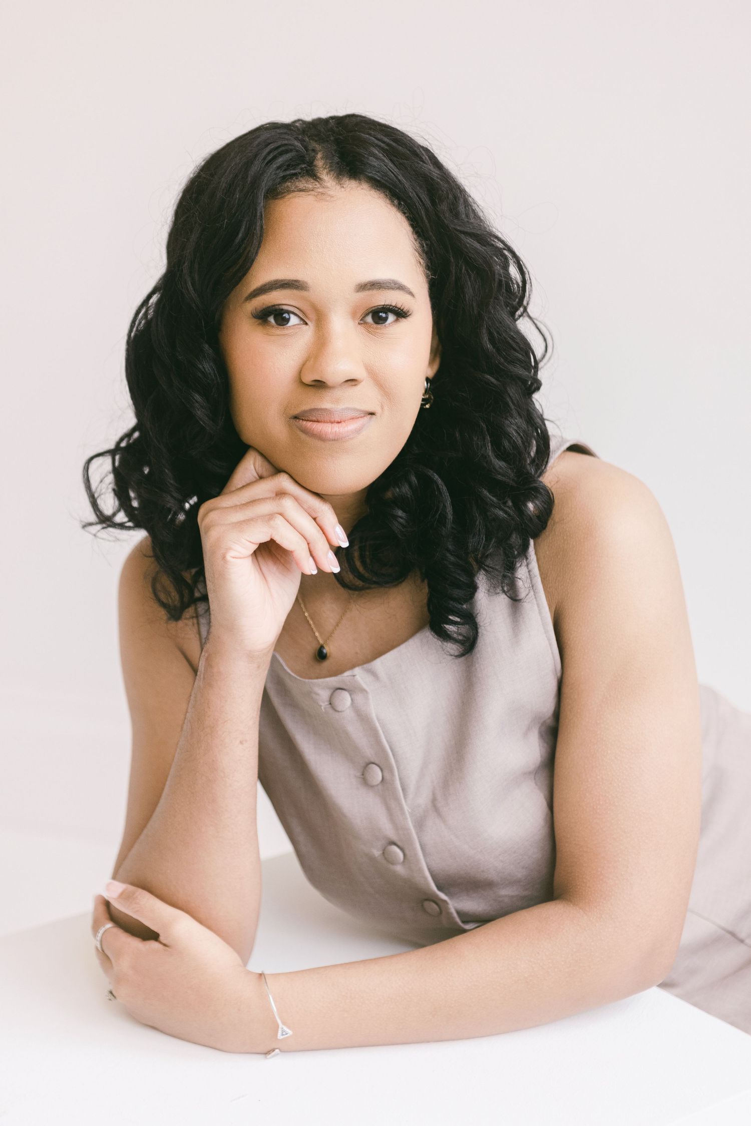 Young woman with curly black hair in a gray sleeveless top poses elegantly with hand on chin against white background.