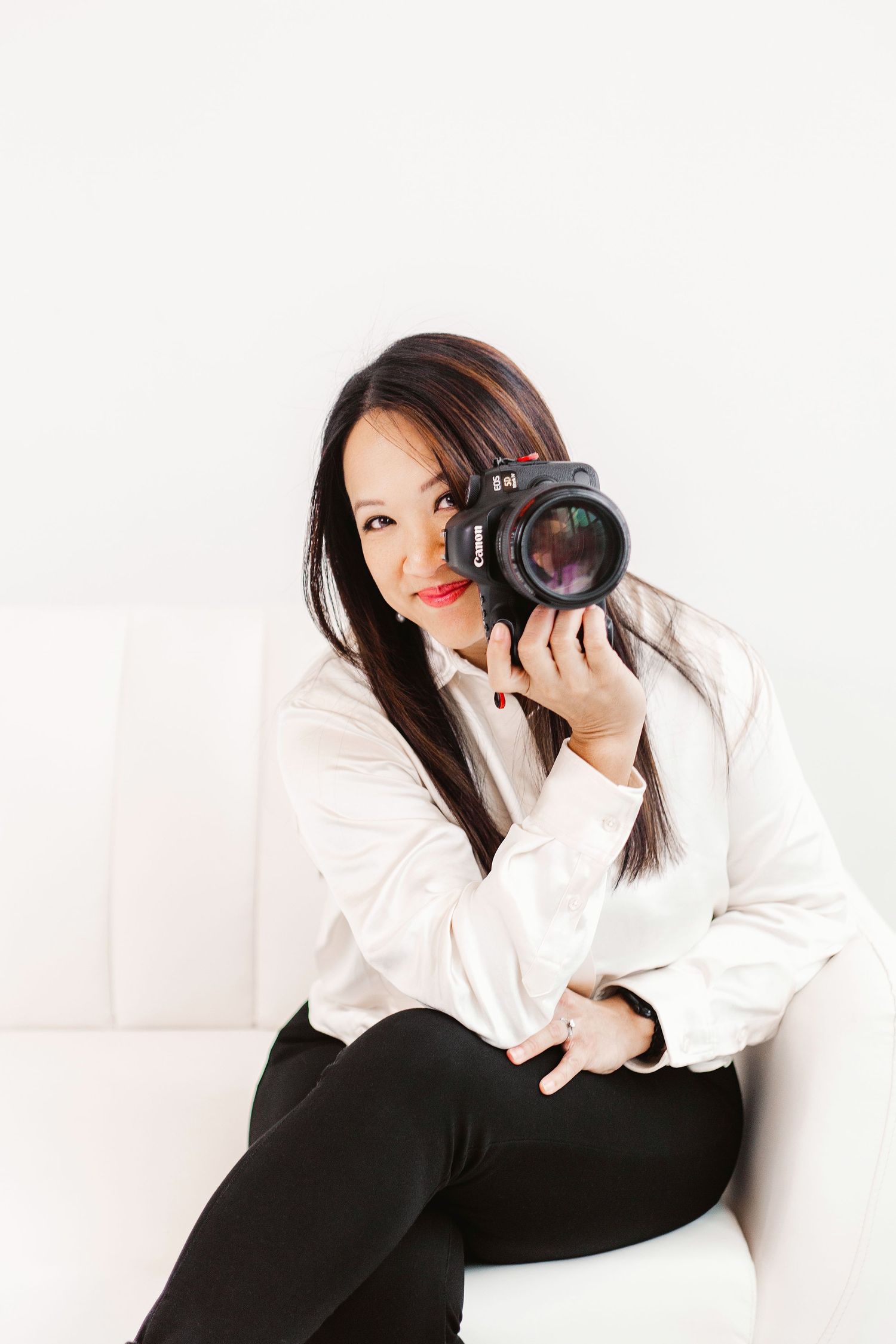 Woman in white shirt sits on sofa holding Canon DSLR camera, smiling at viewer in bright studio setting.