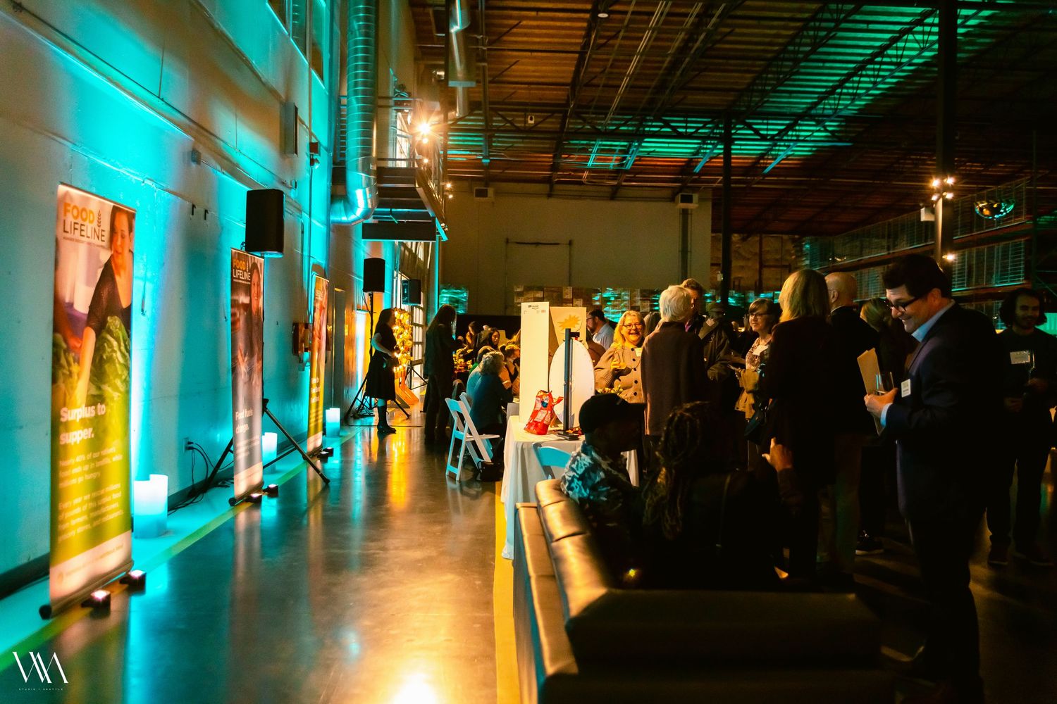 Guests mingle along a gallery wall with teal uplighting at an indoor evening event in a warehouse venue.