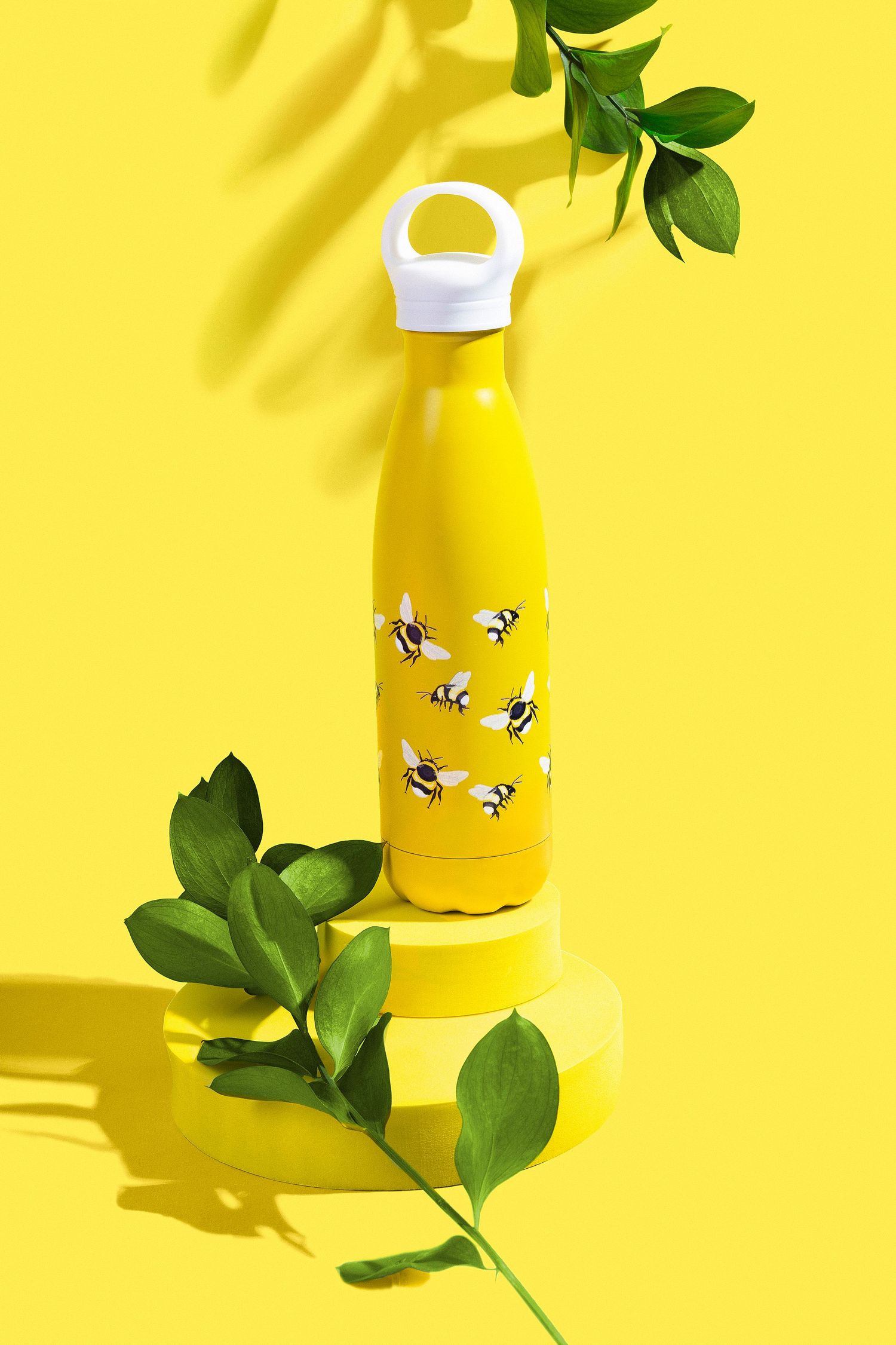 Yellow reusable water bottle with bee print design on yellow podium with green leaves.
