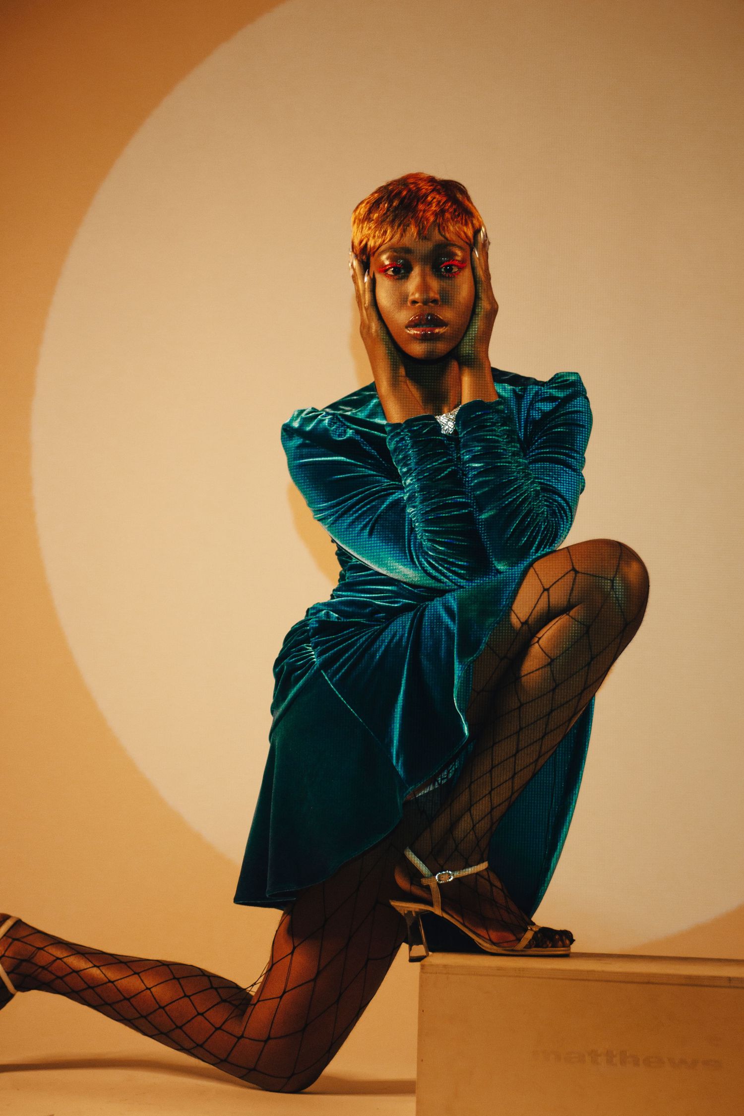A model in a teal velvet dress and fishnet tights poses dramatically on a box against a warm beige background.