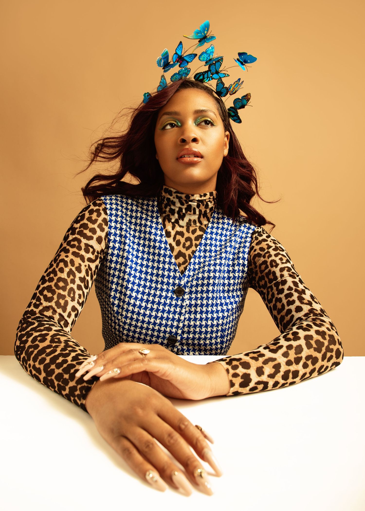 Woman wearing blue butterfly crown, houndstooth vest, and leopard print top poses at white table against tan background.