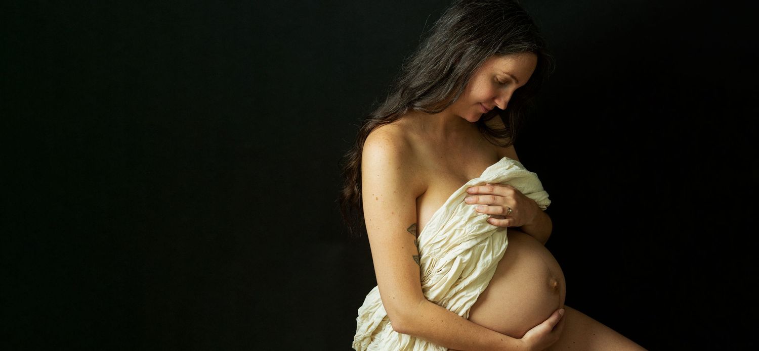 Pregnant woman in white draped fabric cradling her baby bump against a dark background in a maternity portrait.