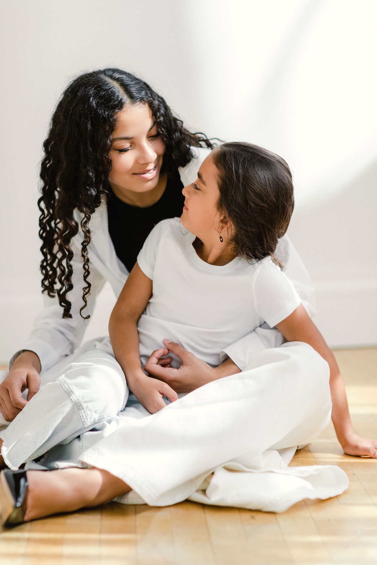 A woman with curly hair and a young girl sit on a hardwood floor, smiling at each other in matching white outfits.