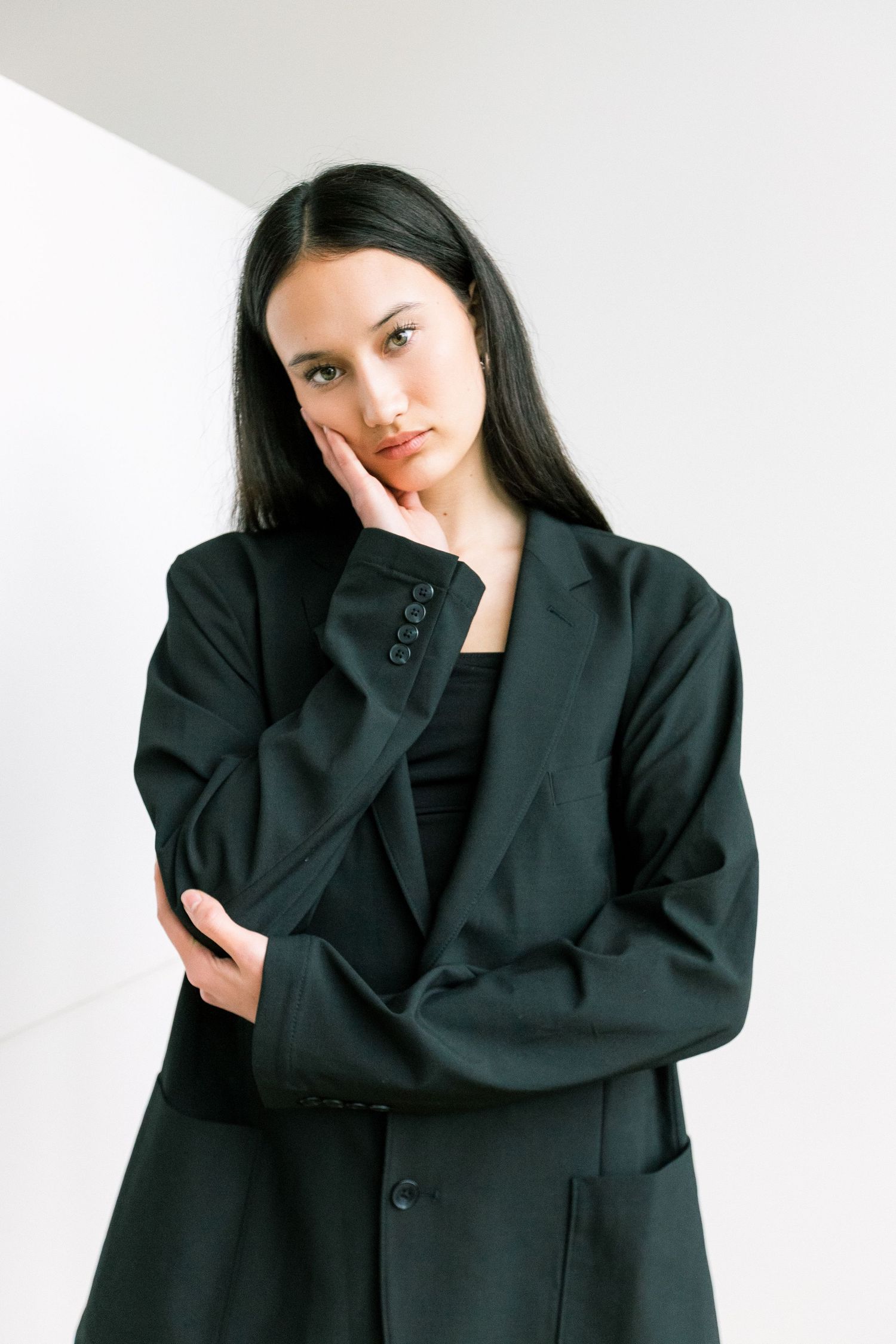 A young woman in an oversized black blazer rests her hand on her cheek against a white wall.