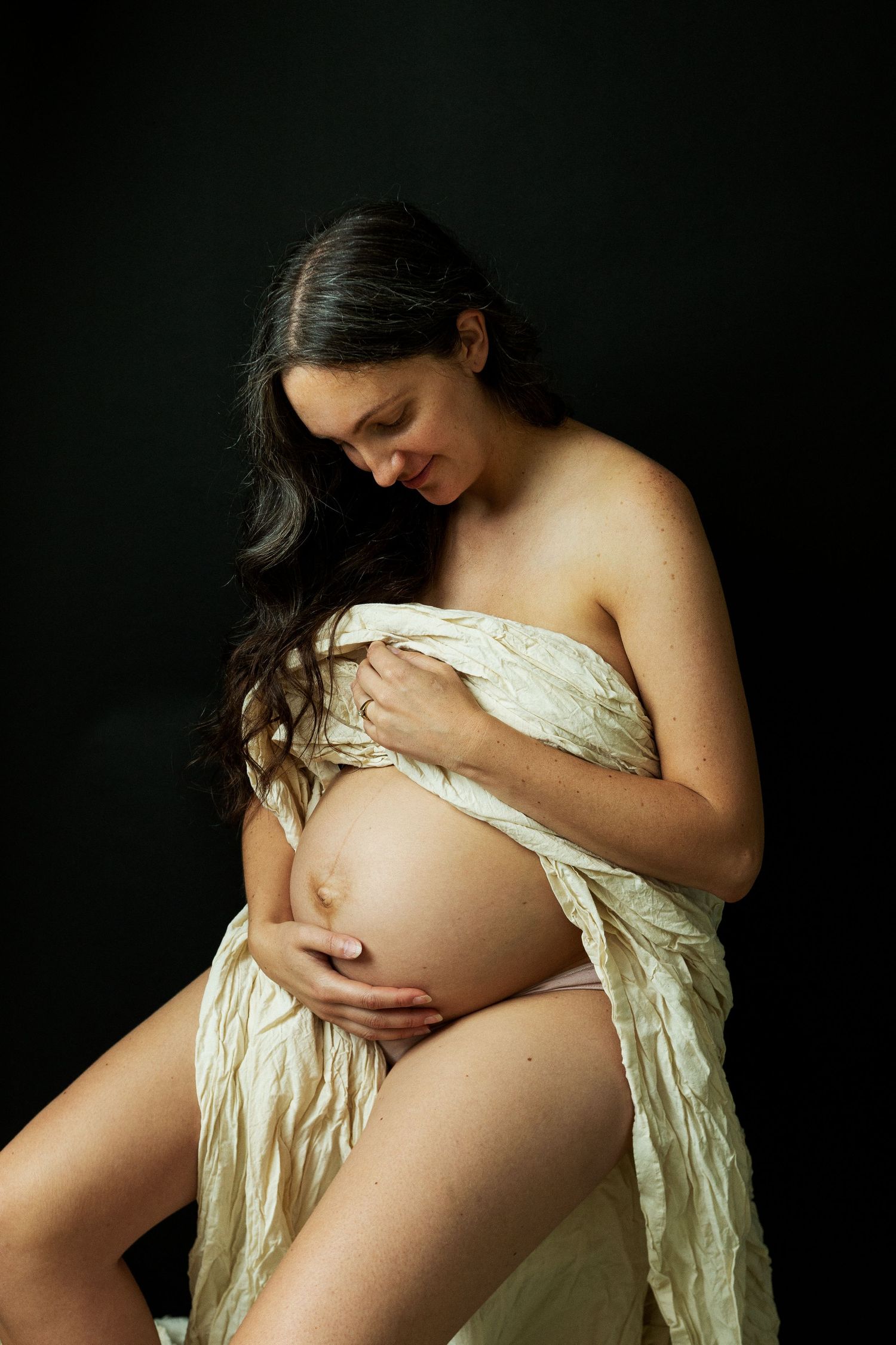 Pregnant woman draped in white fabric cradling her bare belly against a dark black background.