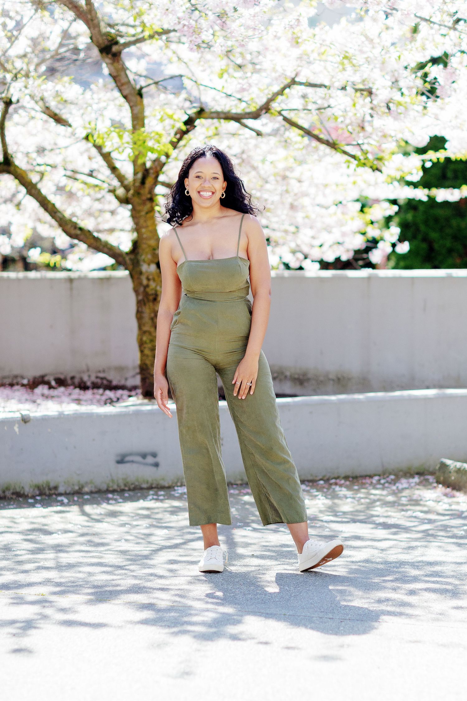 Smiling young woman in olive green jumpsuit and white sneakers posing outdoors near blooming cherry trees.