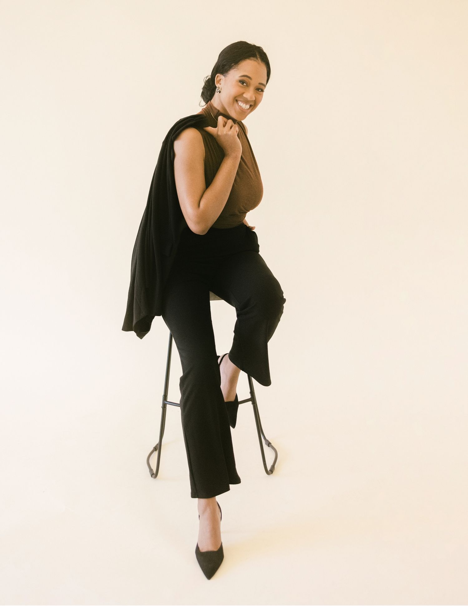 Smiling woman in brown top and black pants sitting on a stool with a blazer draped over her shoulder.