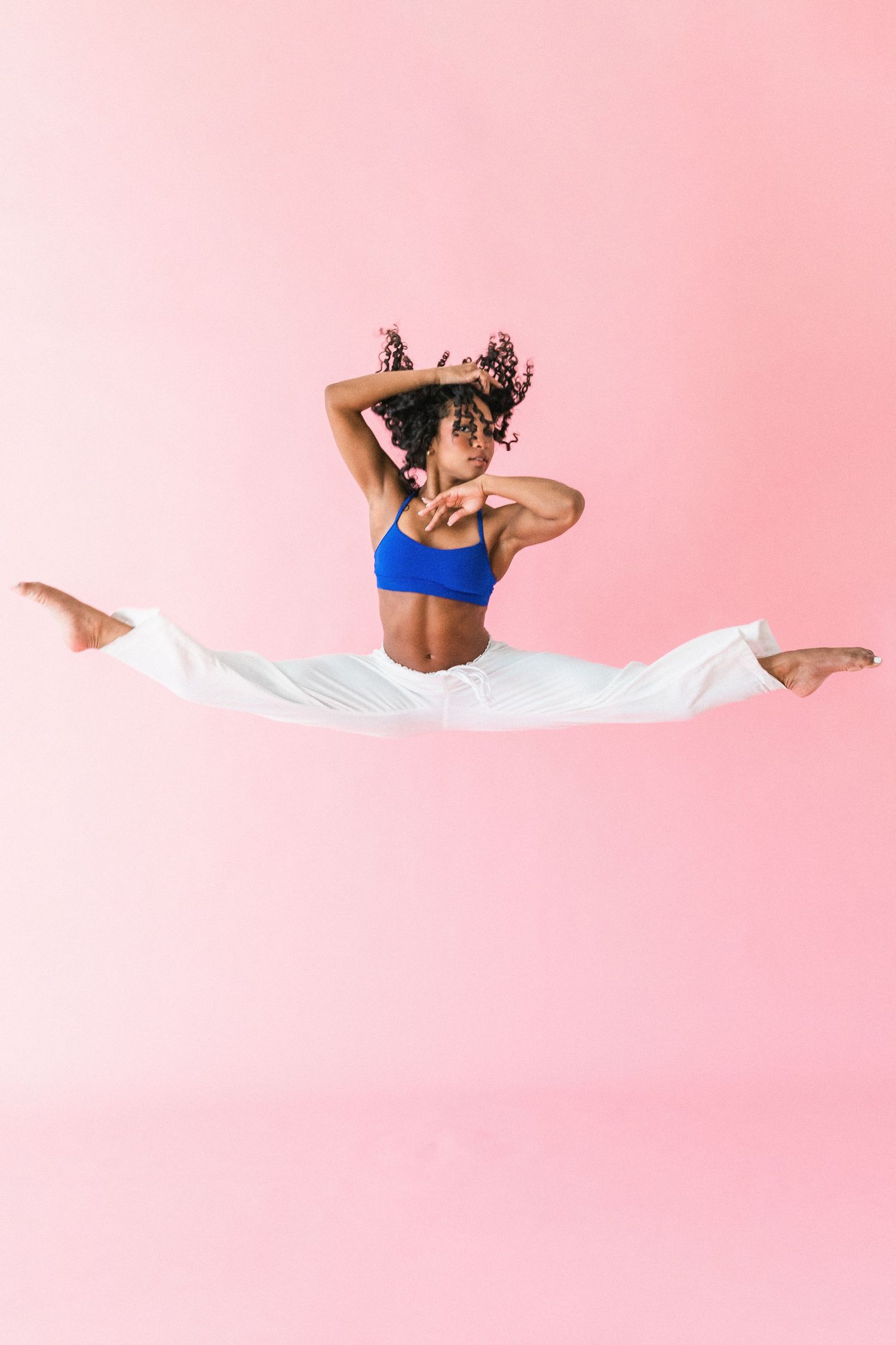A woman in a blue top and white pants performs a mid-air split jump against a pink background.