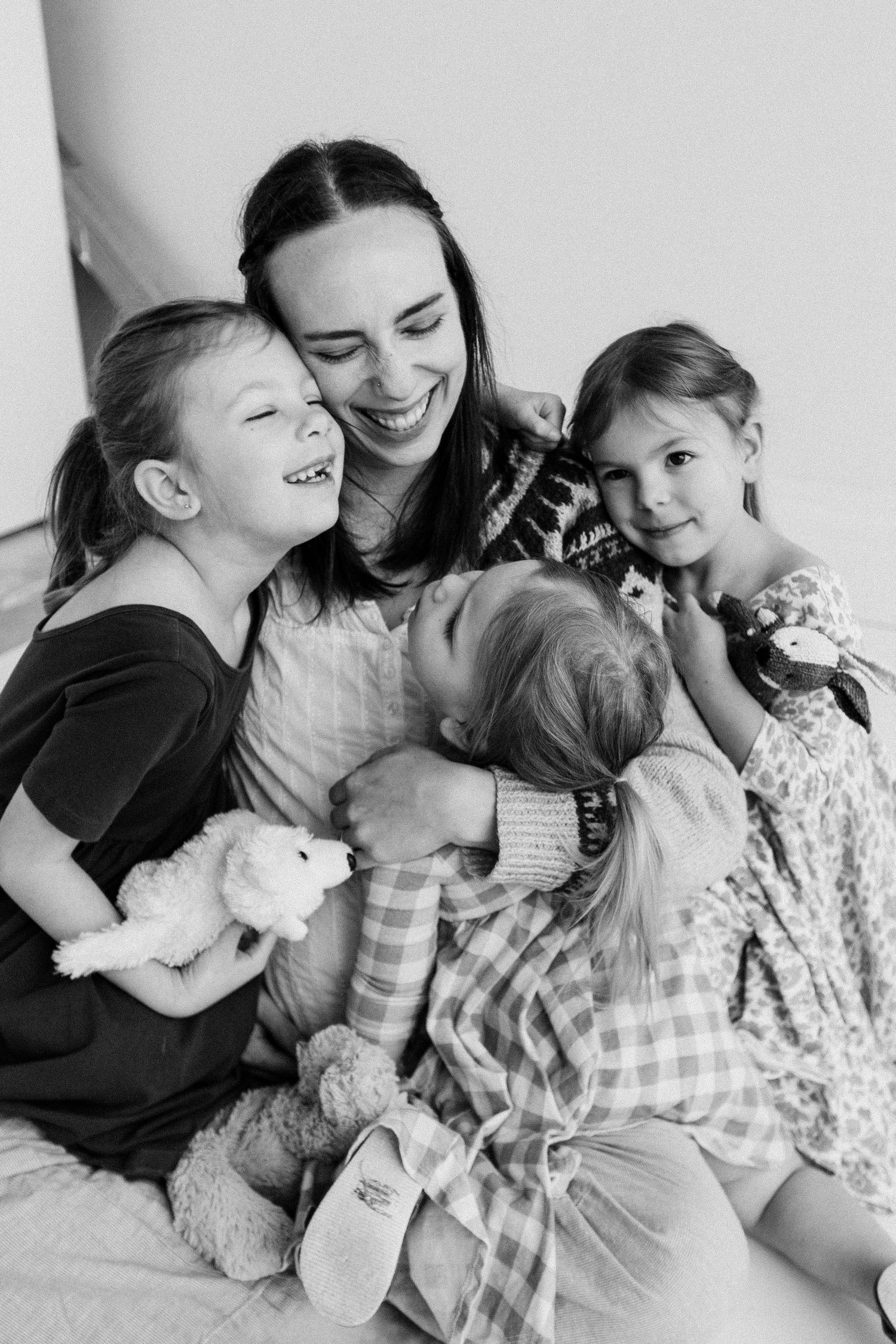 Black and white photo of a smiling mother hugging her three young daughters, all sitting together on a bed.