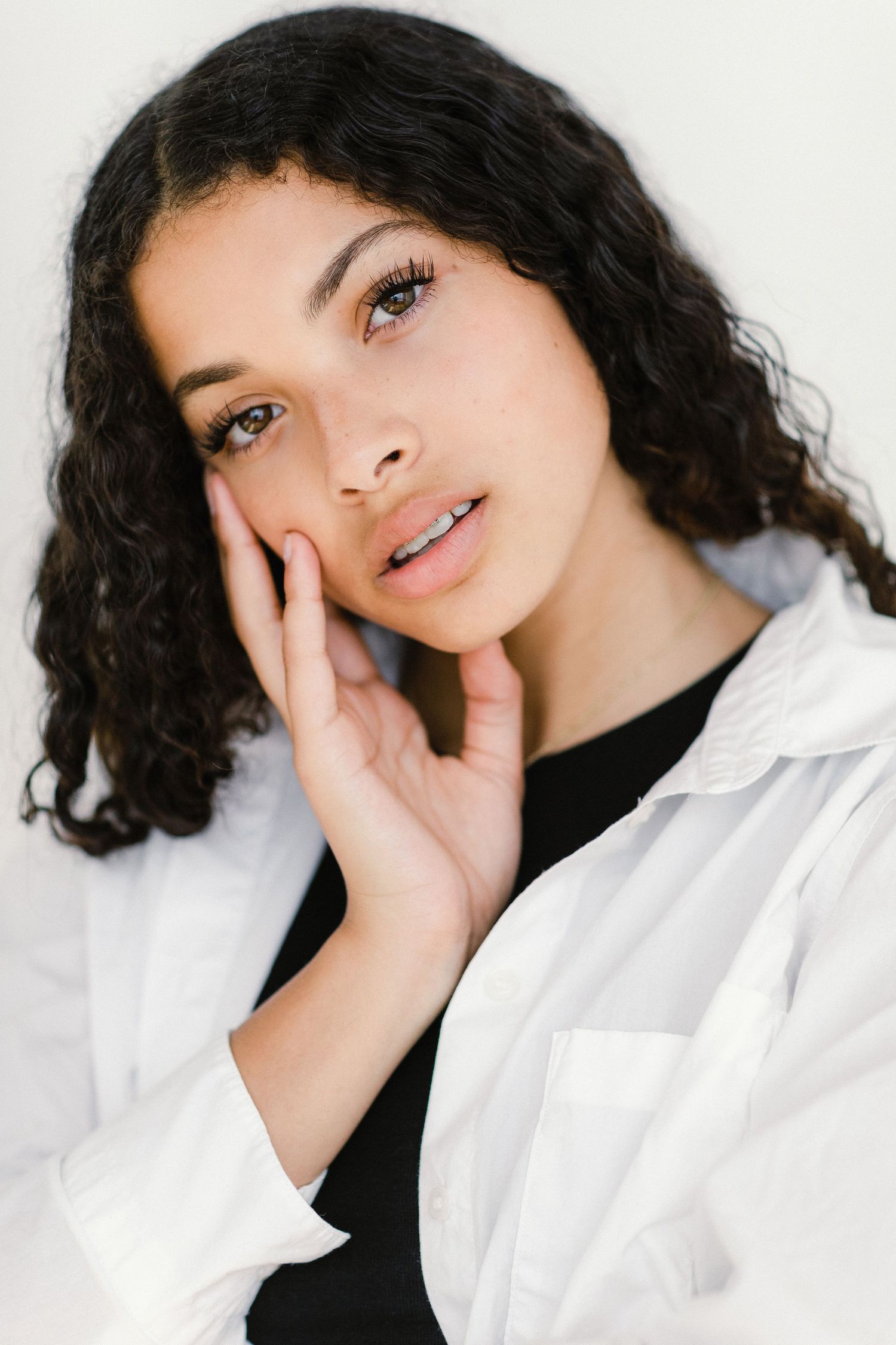 Young woman with curly hair in white shirt touching her face against white background.